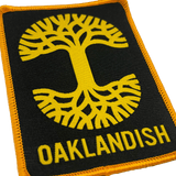 Close-up of black woven iron-on patch with gold Oaklandish tree logo and wordmark.