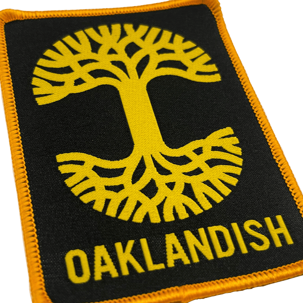 Close-up of black woven iron-on patch with gold Oaklandish tree logo and wordmark.