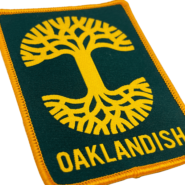 Close-up of forest green woven iron-on patch with gold Oaklandish tree logo and wordmark.