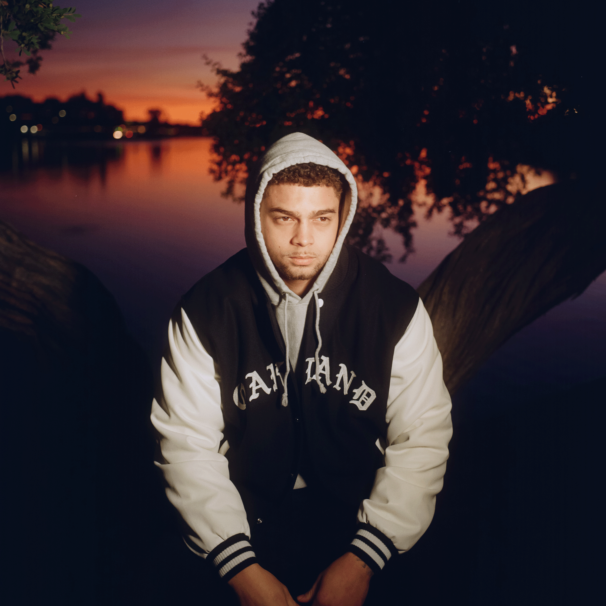 A man sitting beside the lake, facing forward, at sunset wearing a black and white varsity jacket with a large OAKLAND wordmark on the front chest.