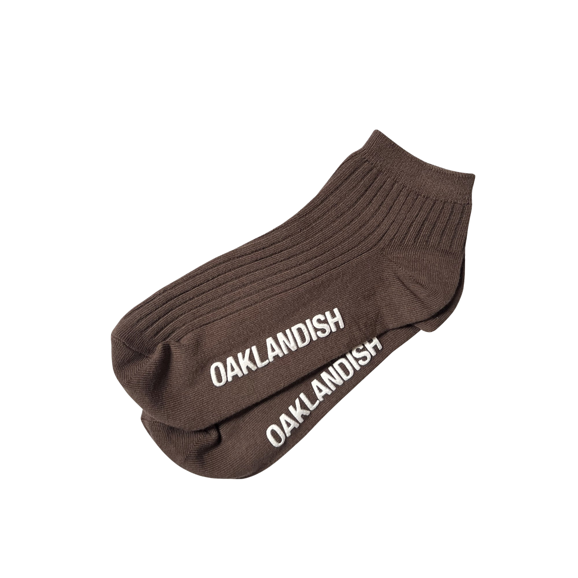 A pair of the dark brown Grip Low Crew Socks by Oaklandish, featuring ribbed fabric and white "OAKLANDISH" text embroidered near the toes. These cotton socks are neatly arranged, overlapping each other on a light gray background.