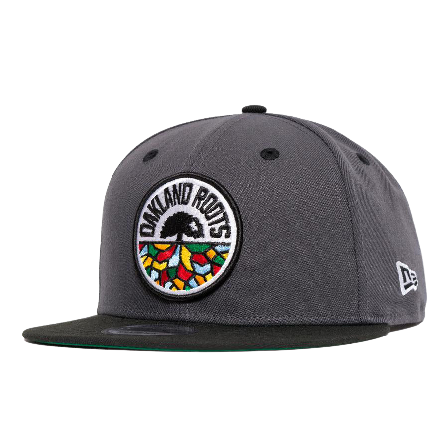 The New Era Oakland Roots SC 59FIFTY Fitted cap, in dark gray, showcases the Oakland Roots Sports Club logo with vibrant embroidery on the front panel. The logo features a tree with roots spreading out in various colors. The New Era logo is stitched onto the side, and the underside of the brim is dark green.
