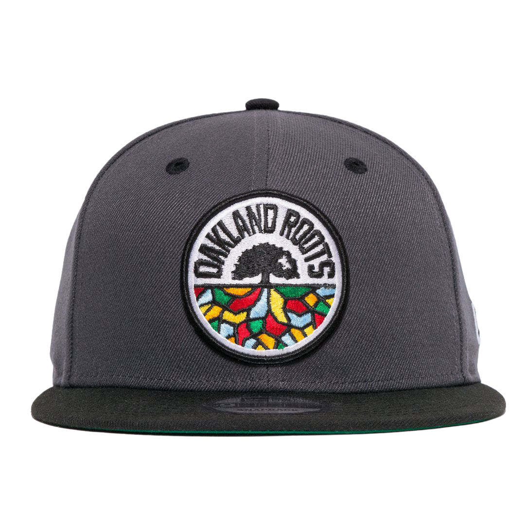 The New Era Oakland Roots SC 59FIFTY Fitted cap, in dark grey, showcases an intricately embroidered logo on the front featuring a black tree with spreading roots. Surrounding the tree is the bold text "OAKLAND ROOTS," accented by colorful shapes in red, yellow, green, and blue that symbolize the foundational pride of this distinguished sports club.