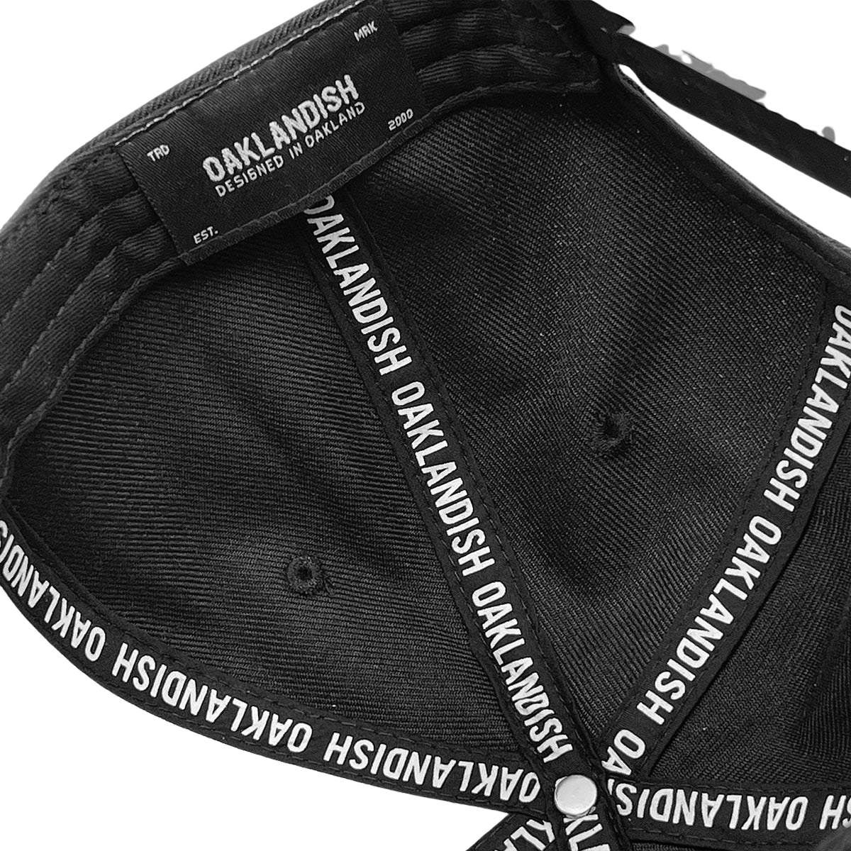 A close-up shot of the inside of the Oaklandish Classic Snapback cap. The fabric on the inside is black, and the edges showcase intricate black and white stitching. The sweatband is adorned with the repeated text "OAKLANDISH." Additionally, a label states "OAKLANDISH DESIGNED IN OAKLAND EST. 2000." This stylish headwear exemplifies Oaklandish's finest craftsmanship.