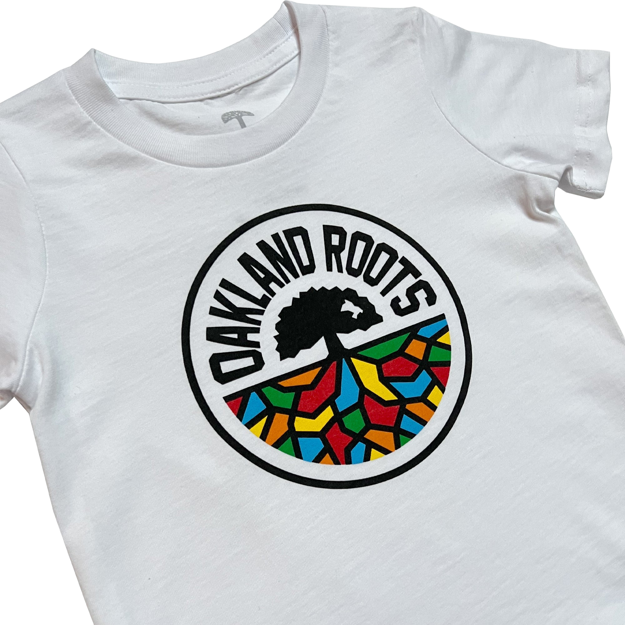 The Toddler Oakland Roots SC Classic Tee displays a circular emblem in the center. The logo, featuring "OAKLAND ROOTS," arches over a silhouetted tree. Below the tree, there is a vibrant mosaic pattern with red, yellow, blue, and green colors that reflects the spirited nature of Oakland Roots SC.