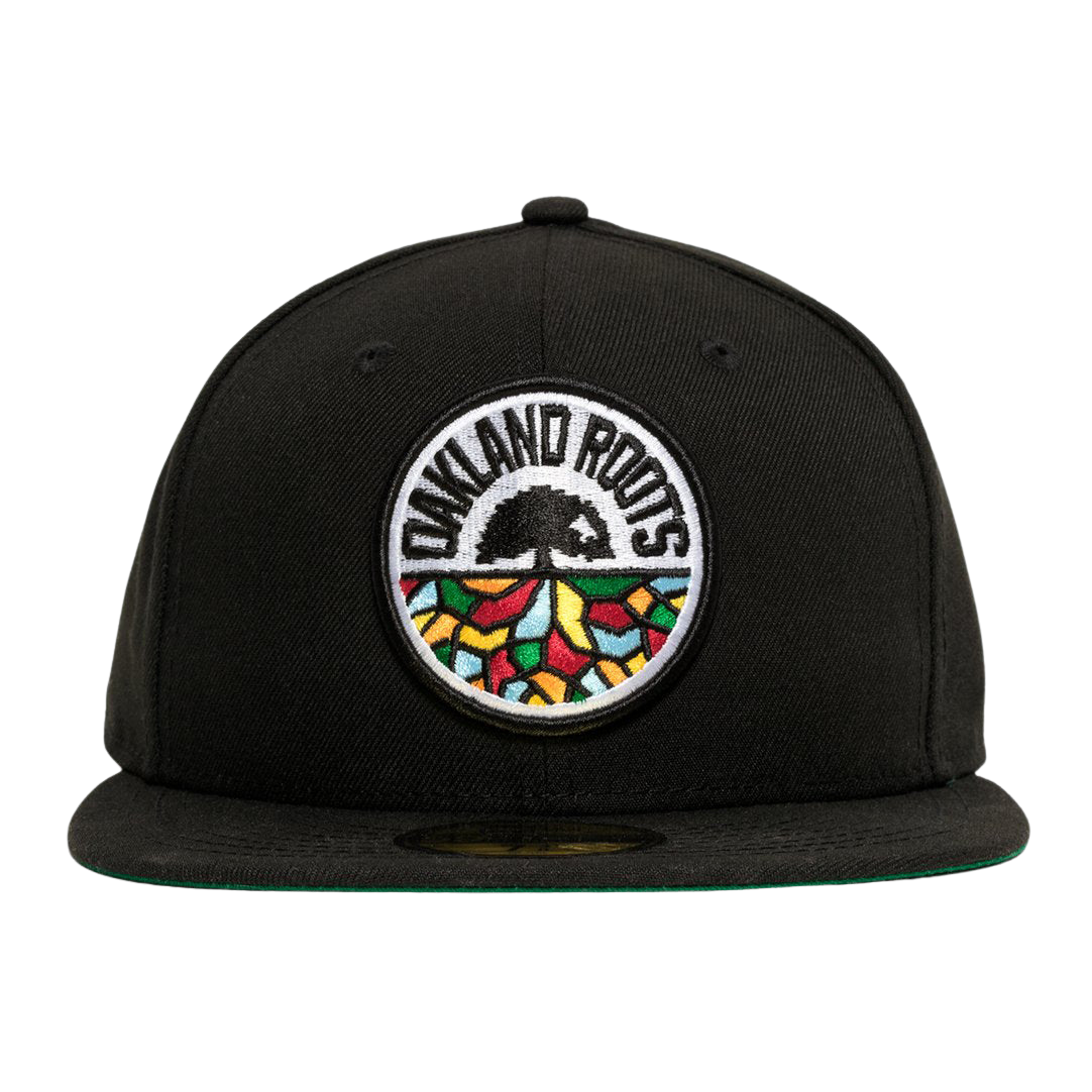 A New Era Oakland Roots SC 59FIFTY Fitted Cap in black, featuring a circular embroidered patch on the front. The patch showcases the words "Oakland Roots" at the top, a black tree in the center, and colorful geometric shapes resembling a mosaic at the bottom. This sports club cap comes with a flat brim and a green underside.