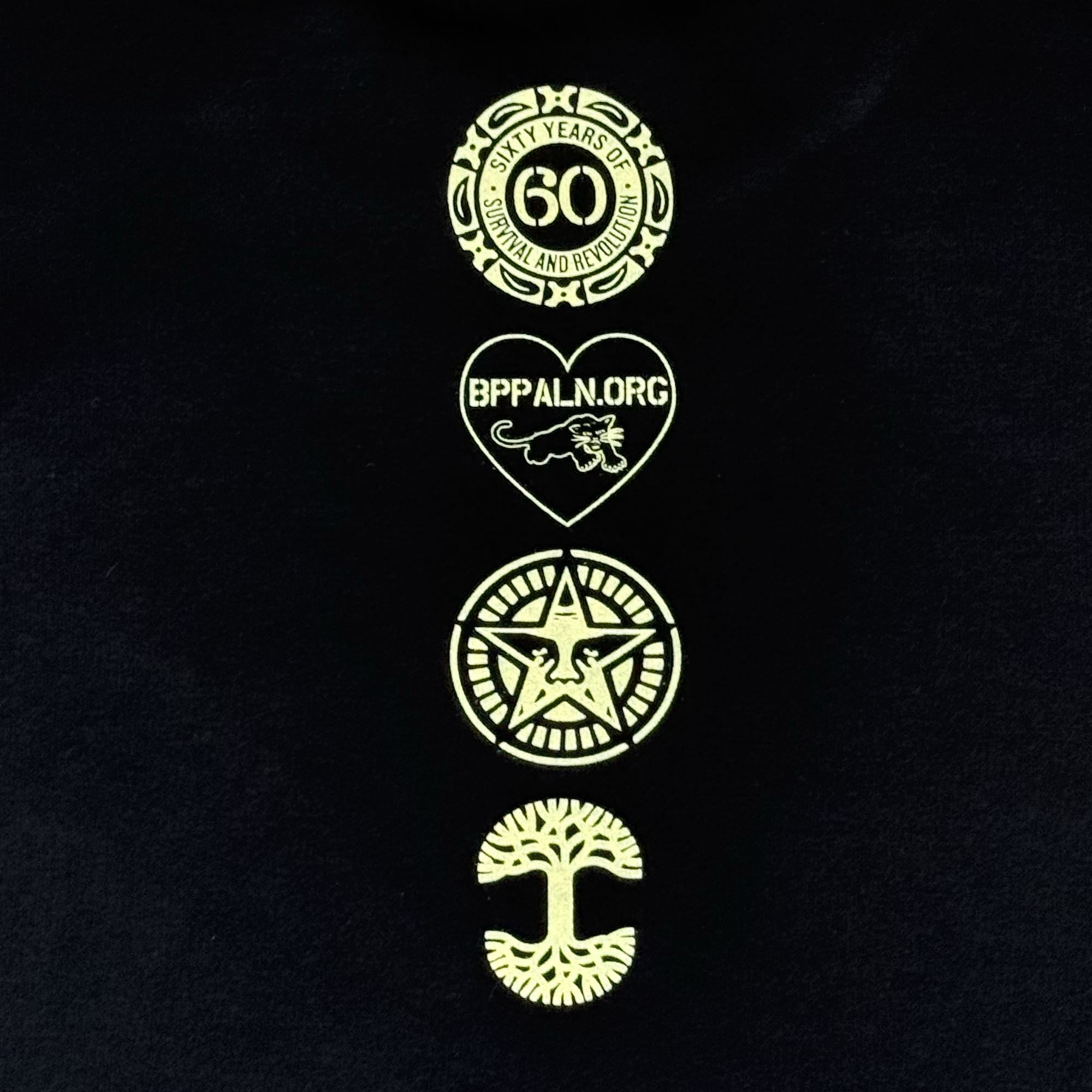 A black backdrop on the Oaklandish Obey x BPPALN 60th Educate to Liberate Crew features four gold logos in a vertical line, celebrating the Black Panther Party’s 60th anniversary with special emblems and a “Sixty Years of Survival and Revolution” badge.