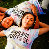 Models wearing Oakland Roots SC Peace, Love, and Oakland tie dye pastel tee on grass. 