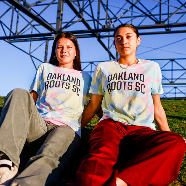 Models wearing Oakland Roots SC Peace, Love, and Oakland tie dye pastel tee sitting on hill of grass.