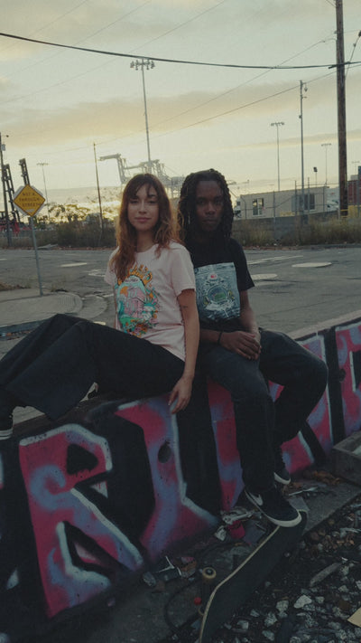 Models outdoors in industrial/port area, female model wearing Oakland Dream tee in pink and male model wearing black Mems tee depicting the Oakland Coliseum.