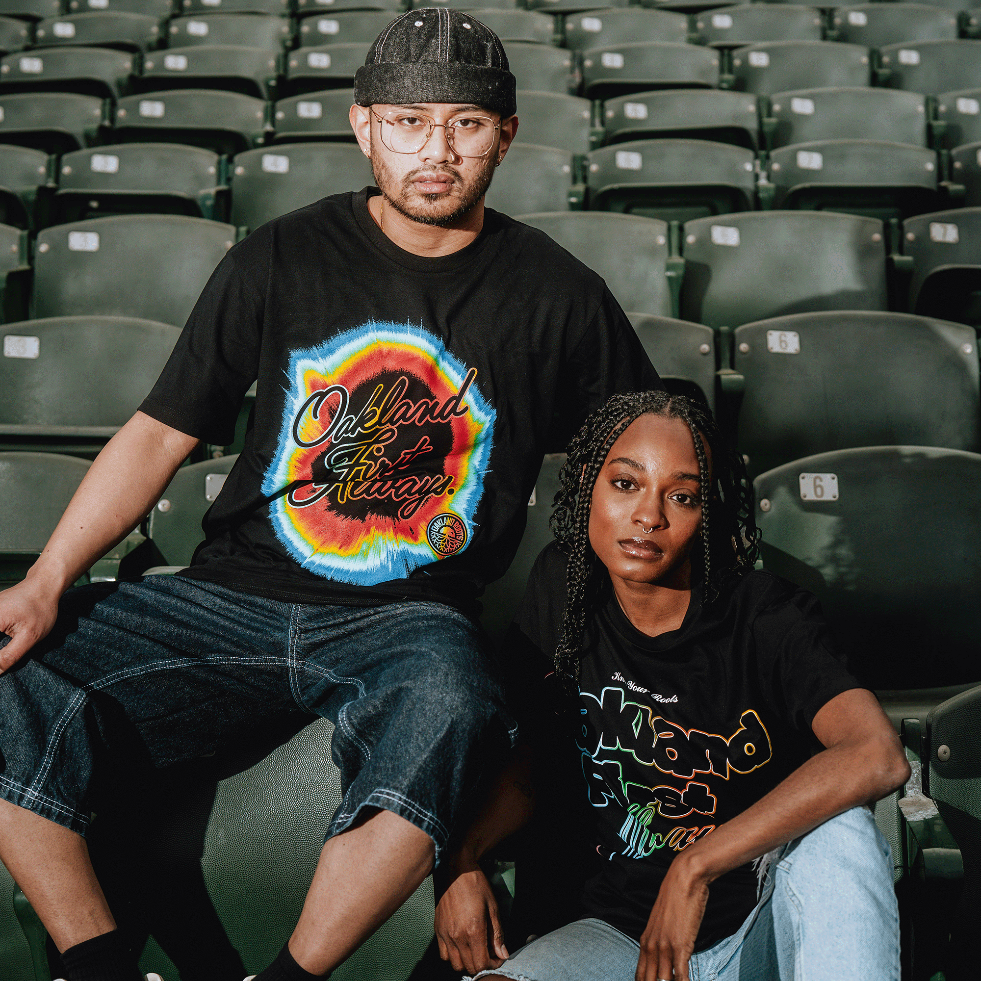 Two fans sit in green stadium seats at an Oakland Roots SC game, both wearing black Oakland Roots SC OFA Rad Roots Tees—one tie-dye, one metallic gold. The man sports glasses and a cap; the woman has braids and rests her arm on her knee.