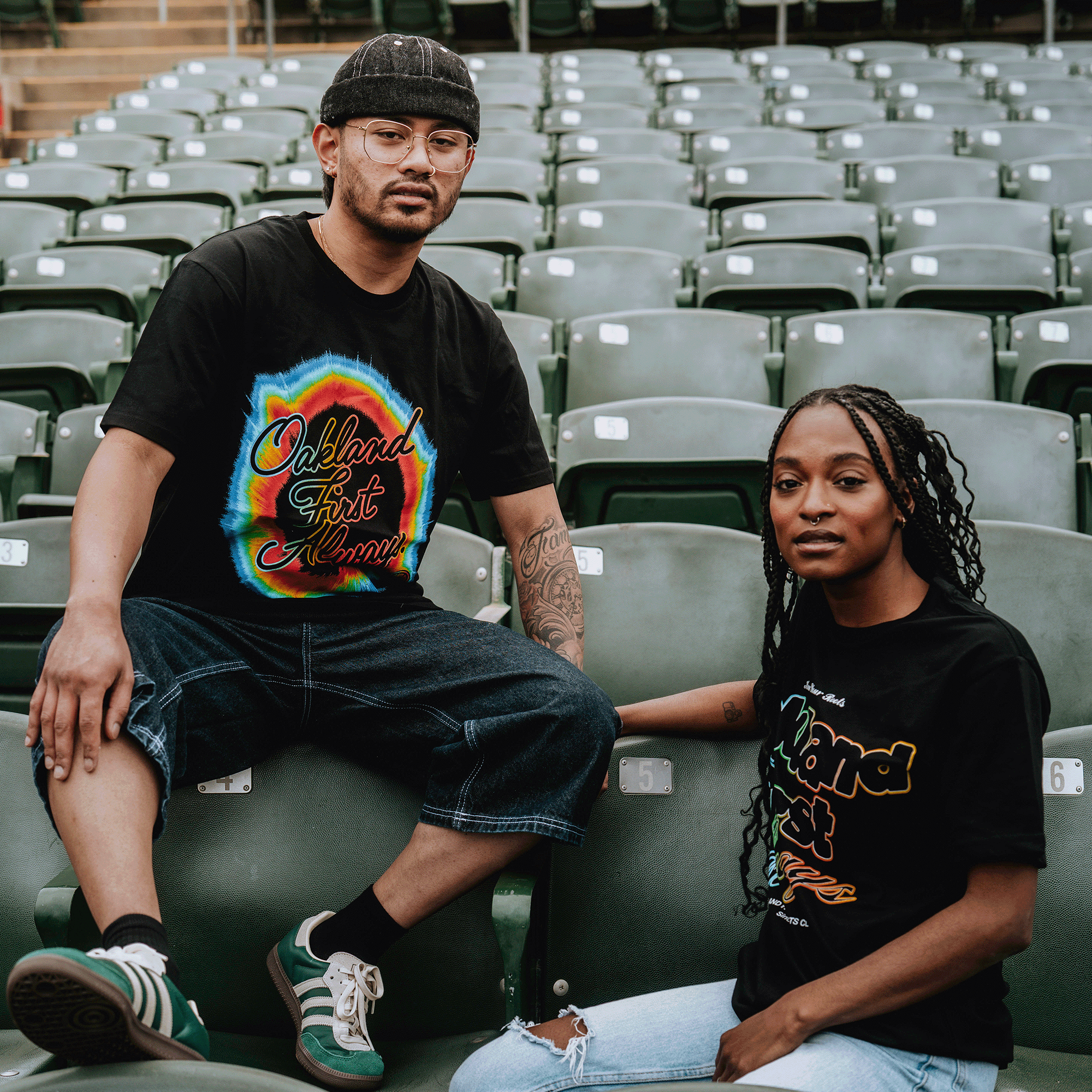 Two people sit among empty green stadium seats, each wearing the Oakland Roots SC OFA Rad Roots Tee. One has glasses and a black cap, the other wears ripped jeans—both proudly show true Oakland Roots spirit to the camera.