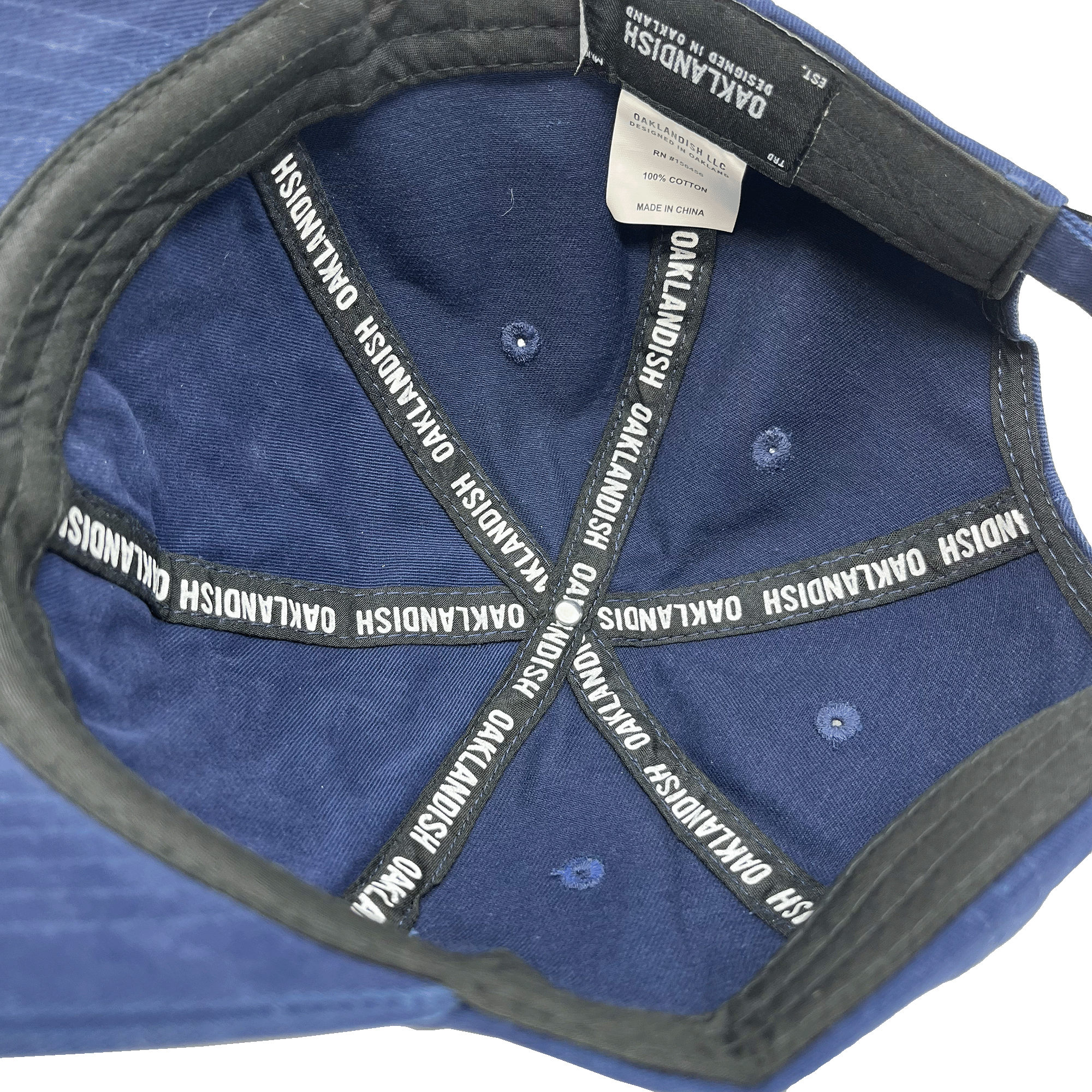 The interior view of the Regal Crest Dad Hat in dark blue from Oaklandish shows eight black straps evenly radiating from the center, each imprinted with the white Oaklandish logo. The black inner band and a white tag are visible, indicating that the hat is made in China from 100% cotton and includes an adjustable strap for a perfect fit.