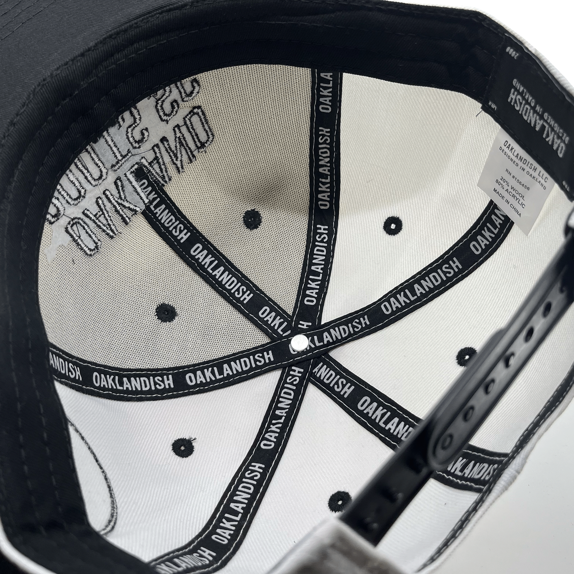 The image showcases the interior of an Oakland Roots SC Arched Text Snapback hat. The inside features white fabric sections printed with "OAKLANDISH" in black, and you can see black ventilation holes and stitching. There is an adjustable strap at the back, along with a white label bearing care instructions.