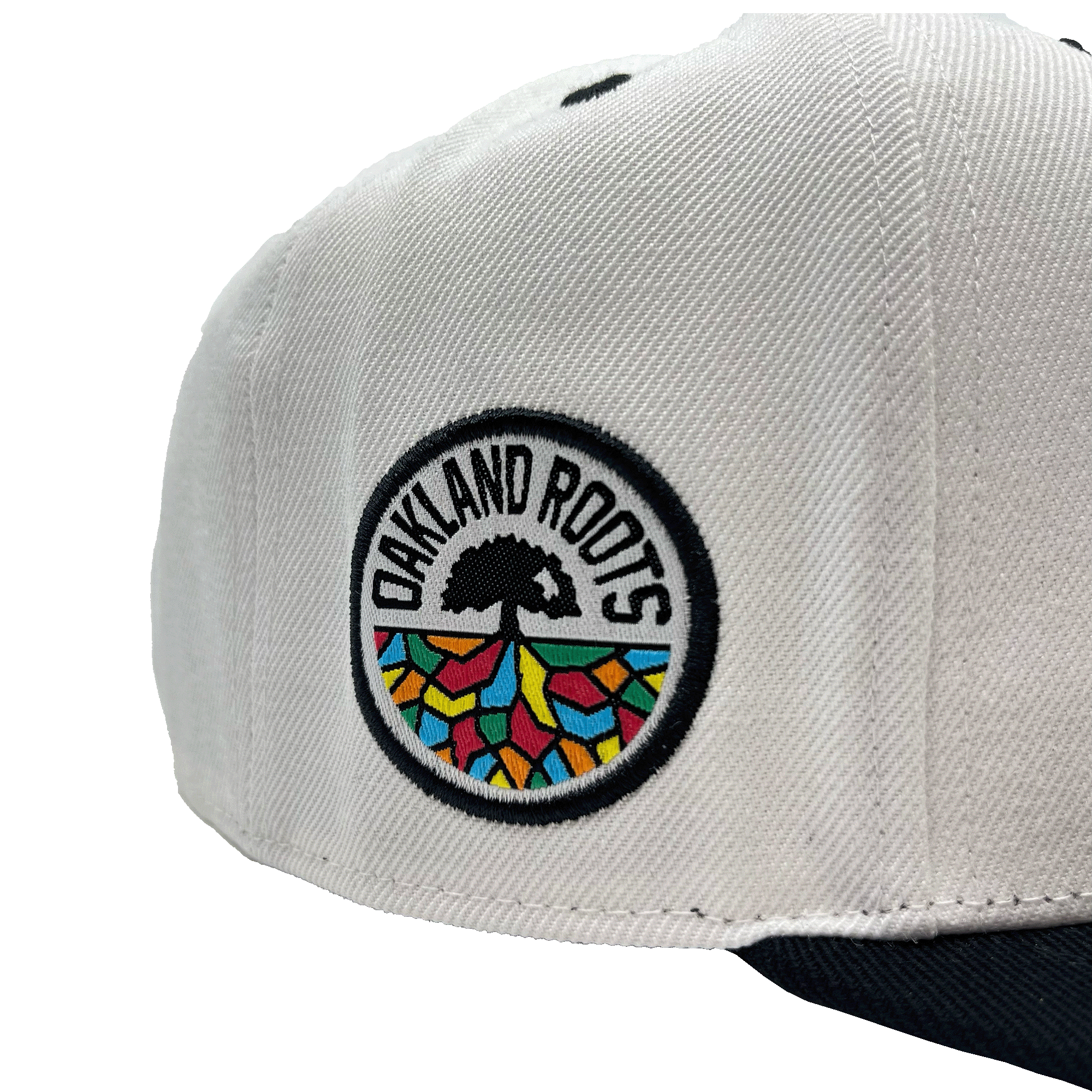 Close-up of the Oakland Roots SC Arched Text Snapback, a white hat featuring a colorful round patch on the side. The patch displays a tree with black roots and rainbow-colored geometric shapes below. The text "OAKLAND ROOTS" encircles the tree symbol in black capital letters, reminiscent of Oaklandish style. The bill of the cap is dark.