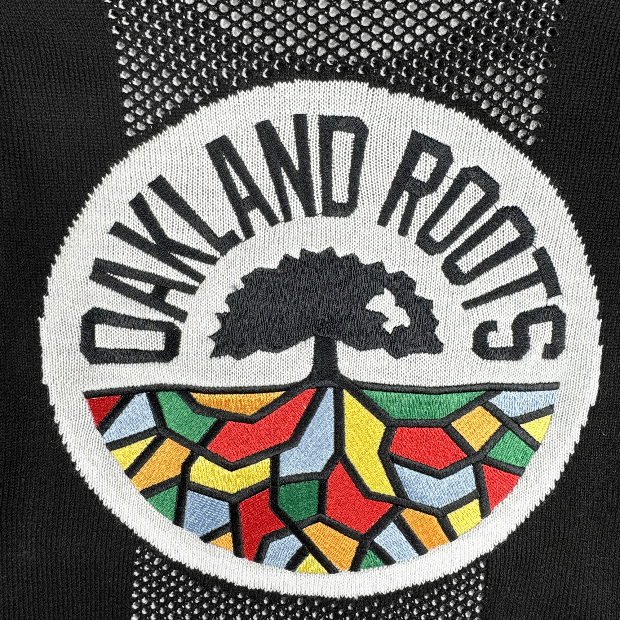 The Oakland Roots SC Clubhouse Sweater features a circular embroidered patch with "OAKLAND ROOTS" above a black tree and vibrant geometric sections in red, yellow, green, and blue, reflecting the spirit of Oakland Roots SC.