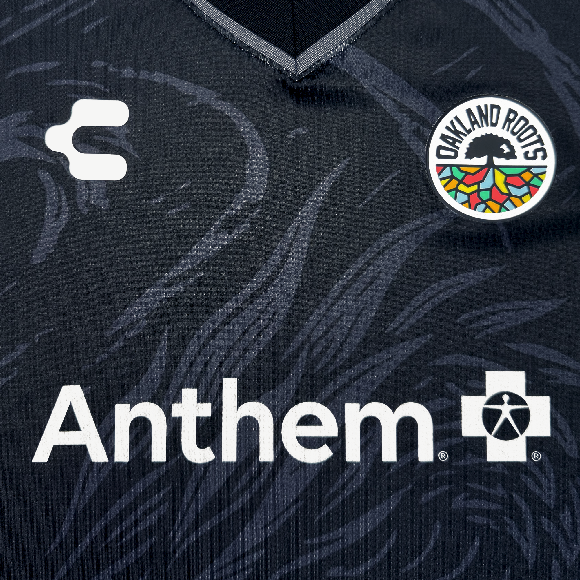 Close-up of the Unisex Oak-ness Awakens Kit by Oakland Roots SC, a dark jersey with an abstract Lake Merritt and Oakland folklore pattern, round team badge, white “C” logo on the left, and bold white Anthem+ sponsor text below.