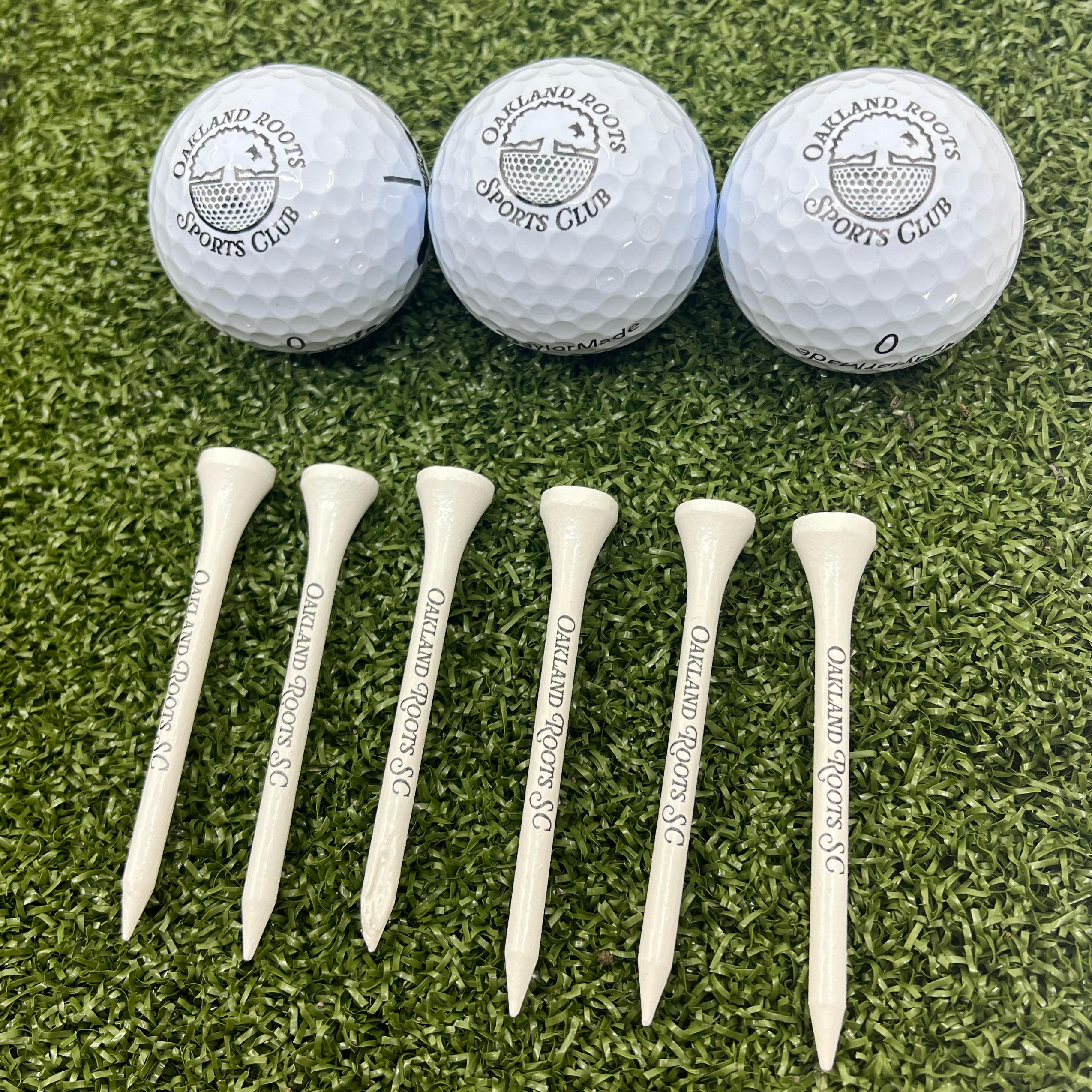 The "Heritage Tournament Pack" from Oakland Roots SC is beautifully displayed with three golf balls featuring the club's logo, all neatly arranged on artificial grass as a symbol of community spirit. Accompanying them are seven white tees printed with "Oakland Roots SC" in black, emphasizing the club’s commitment to social good.