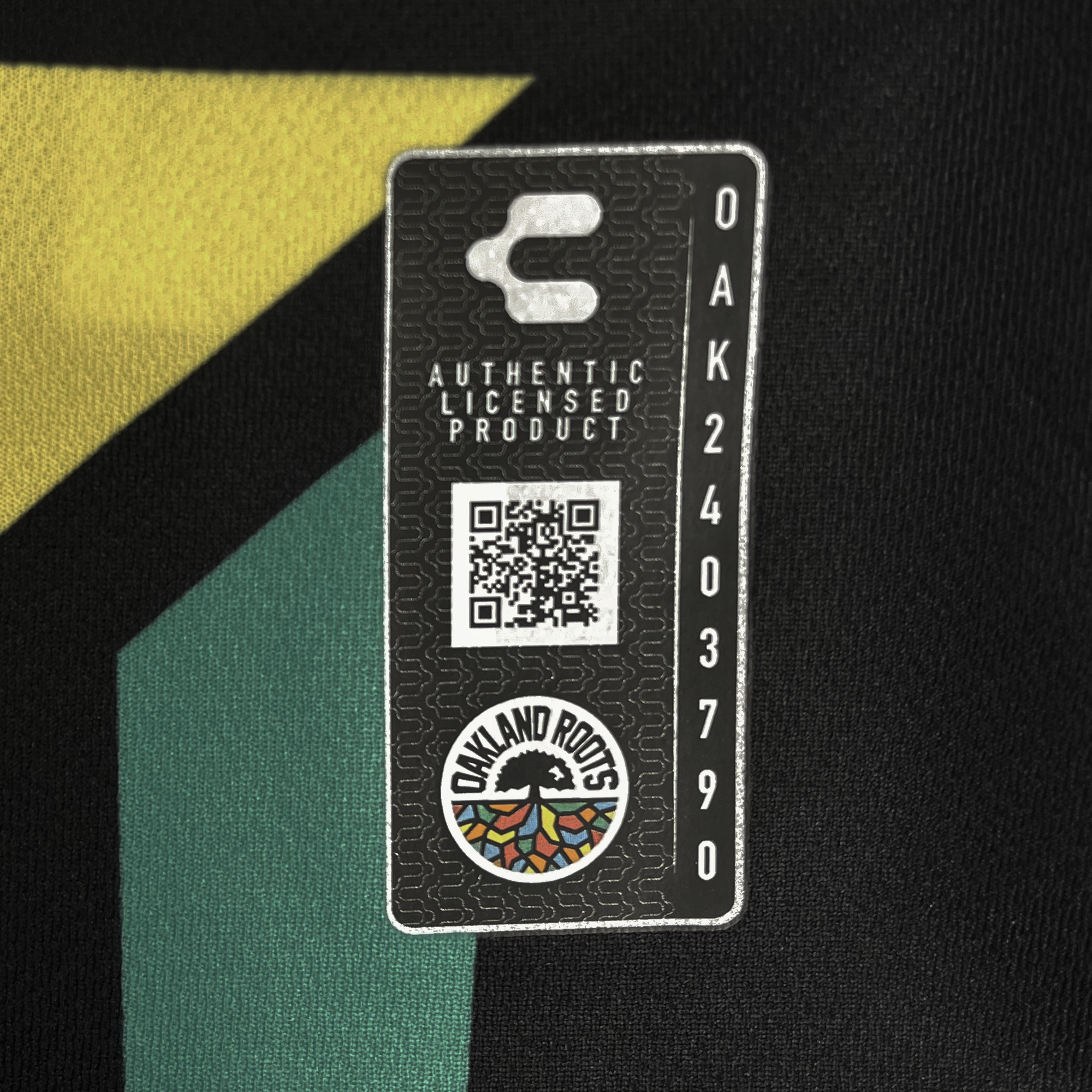 A black fabric tag displays a QR code labeled "AUTHENTIC LICENSED PRODUCT" with "OAK2403790" beneath, surrounded by the "OAKLAND ROOTS" logo. The logo shows a vibrant tree over yellow and green geometrics, capturing inclusivity. Product: Unisex Oakland Roots SC 2025 First Kit ft. Anthem Blue Cross.