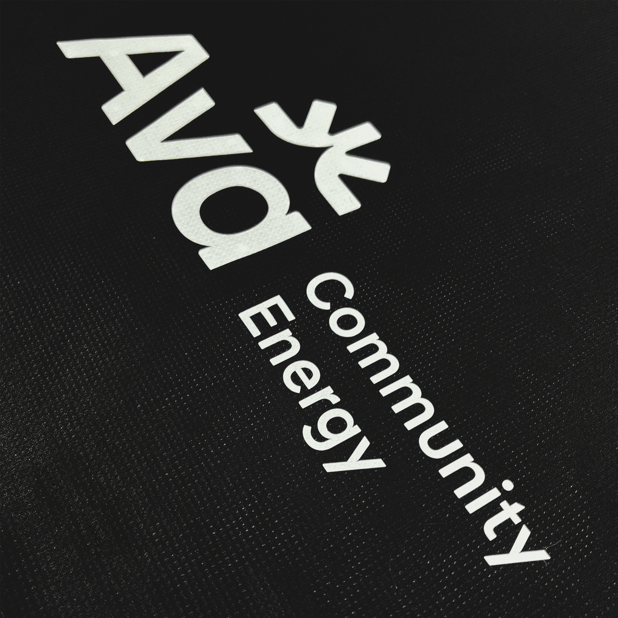 The black background features "Ava Community Energy" in white, with "Ava" incorporating a tree design of geometric shapes and a unity circle. It reflects sustainability and community, inspired by Oakland Roots SC's spirit of renewal and collective energy, as seen in their 2025 first kit with Anthem Blue Cross.