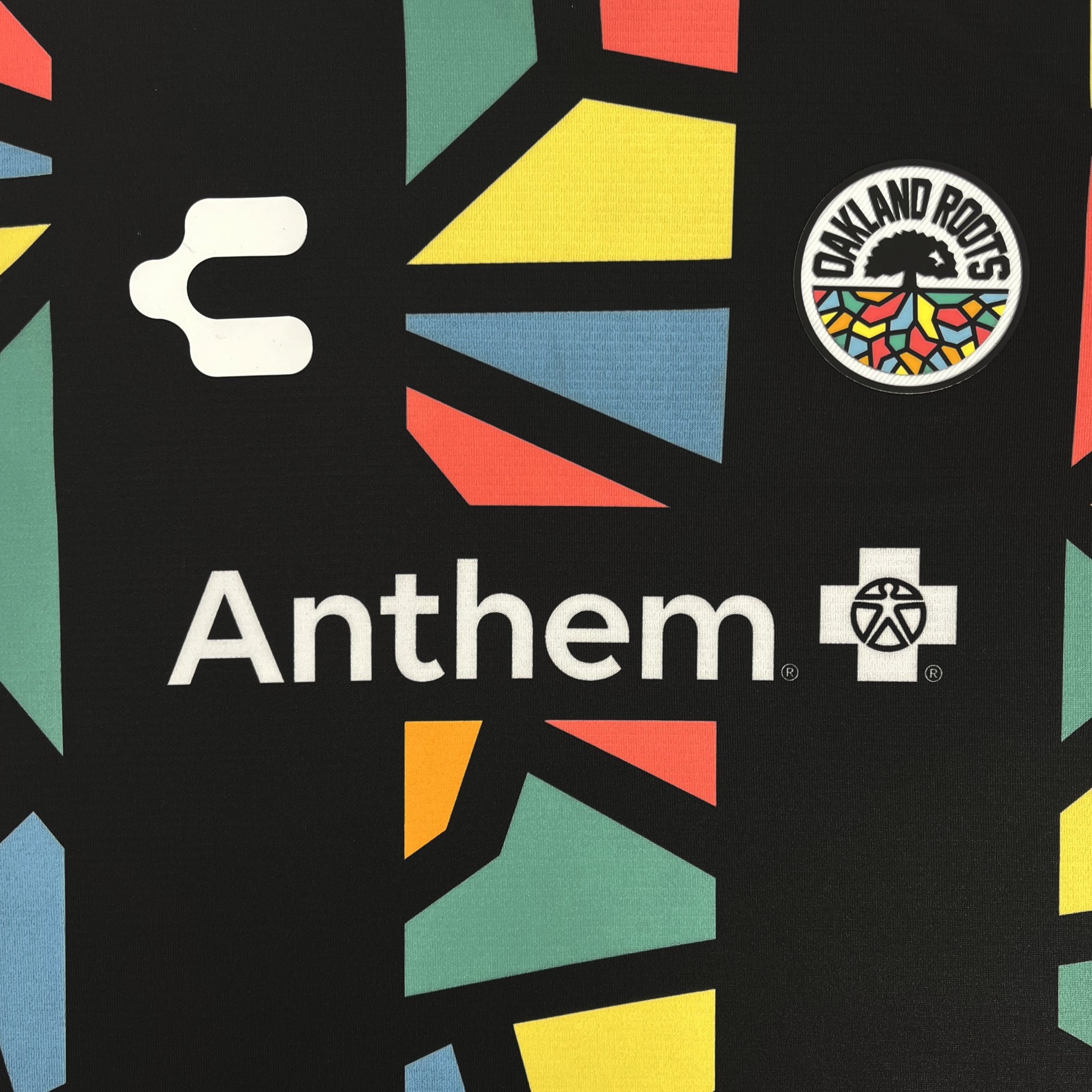 The Custom Unisex Oakland Roots SC 2025 First Kit from Oakland Roots SC features a black jersey with colorful red, green, yellow, and blue geometric shapes. The "Oakland Roots" logo with tree and rainbow shades is on the upper right; "Anthem +" and a patterned globe icon are at the center.
