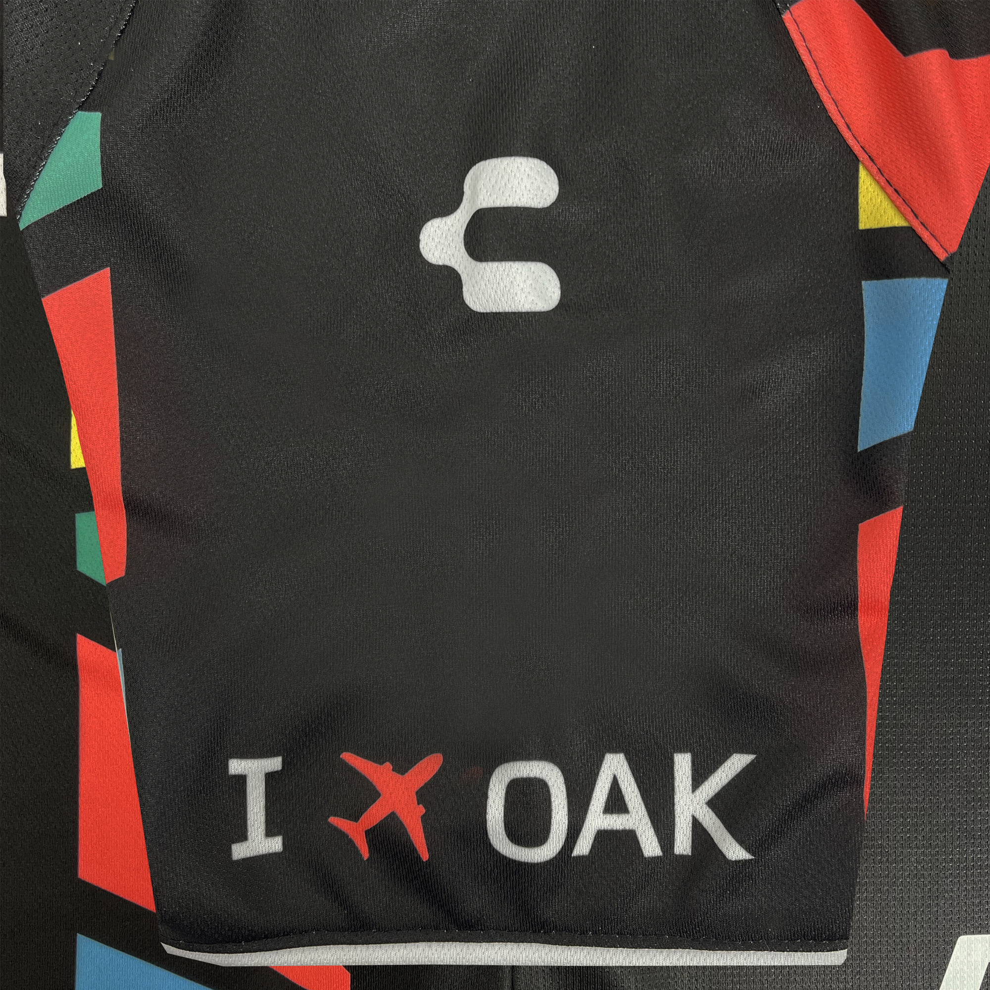 A close-up of the Custom Unisex Oakland Roots SC 2025 First Kit shows a black fabric with vibrant geometric shapes in red, green, blue, and yellow on the sides. The top features a white abstract logo with "I," a red airplane symbol, and "OAK" in white at the bottom for Oakland Roots SC.