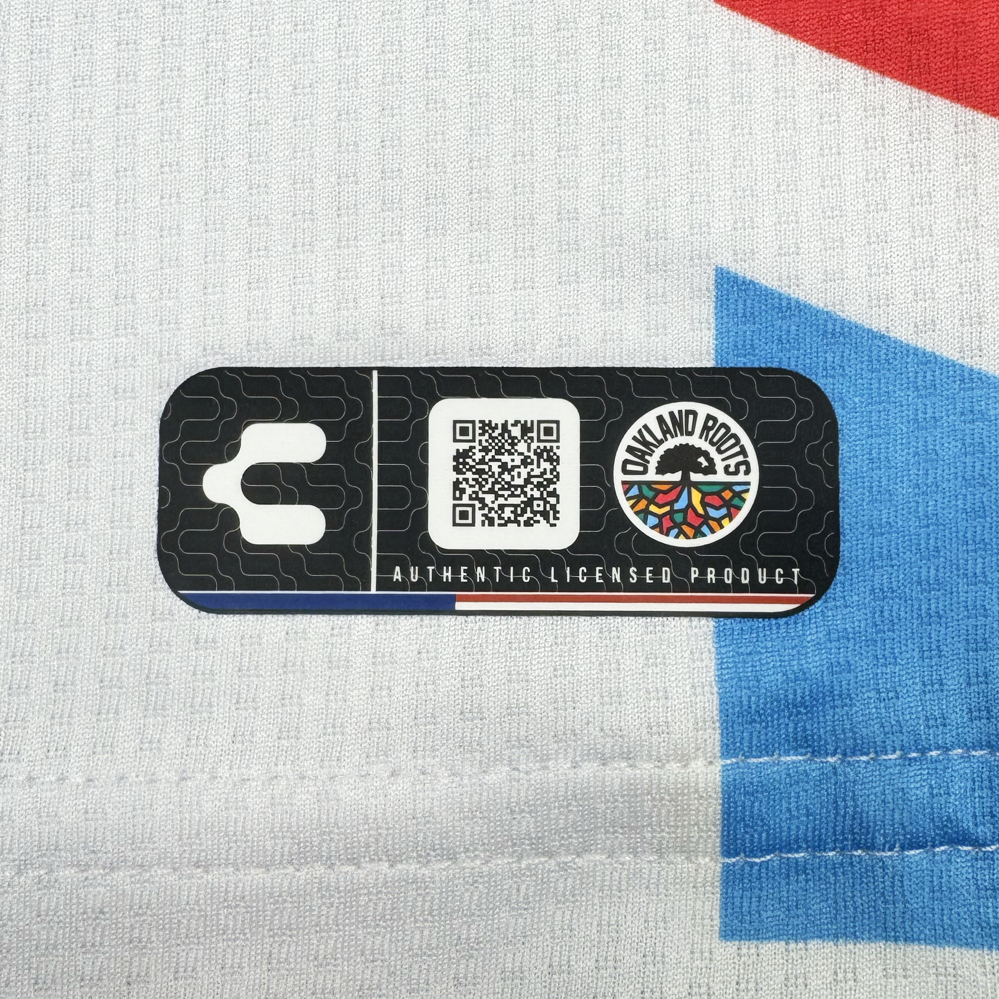 A black rectangular label with a QR code, the "Oakland Roots SC" logo, and "Authentic Licensed Product" is sewn onto the white Custom Unisex Mosaic in Motion Kit, featuring blue and red geometric shapes and visible white stitching at the bottom.