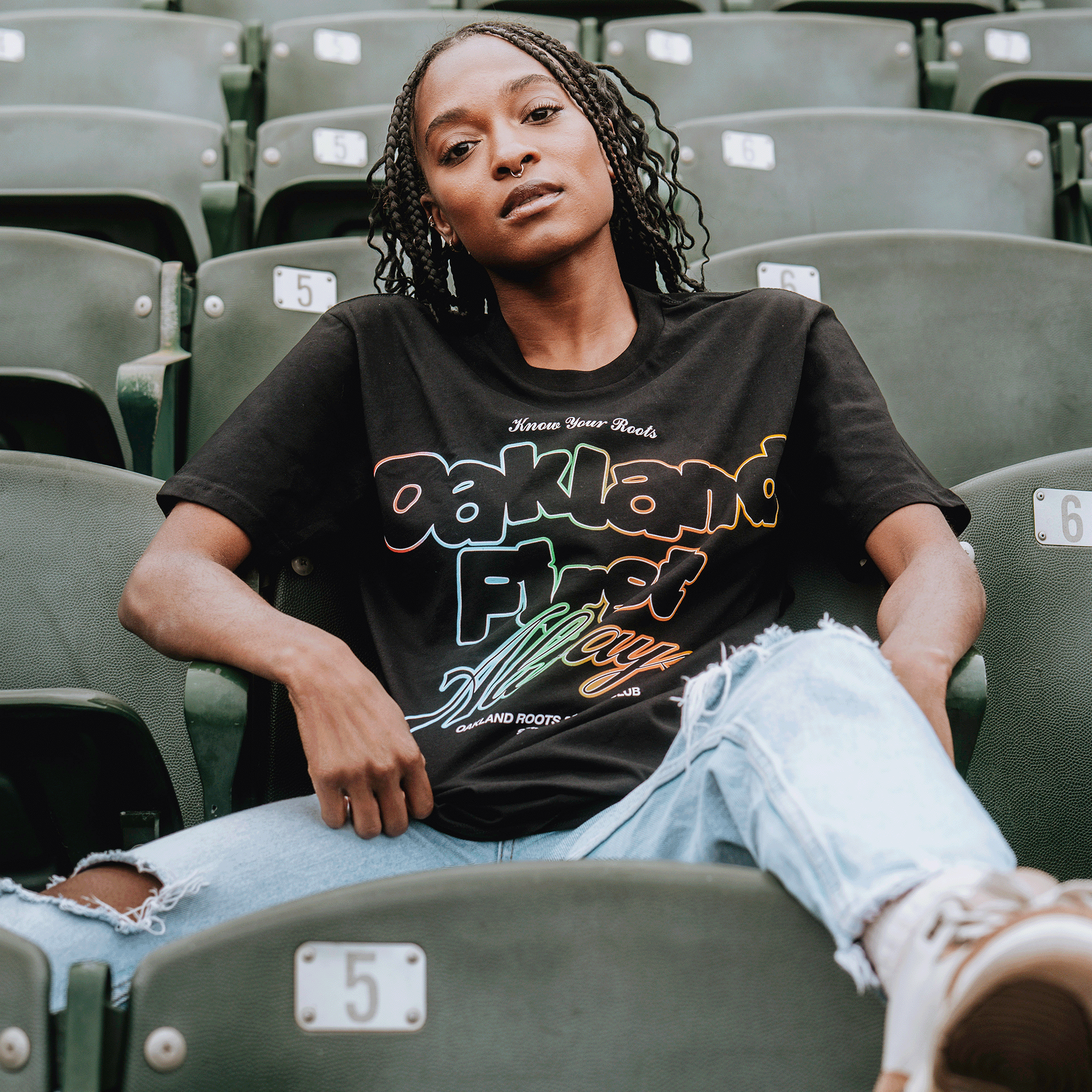 A woman with braids sits on green stadium seats, one leg up, wearing the Oakland Roots SC OFA Different Tee and ripped light blue jeans. She gazes confidently at the camera, displaying true Oakland pride amid white-plaque seats.