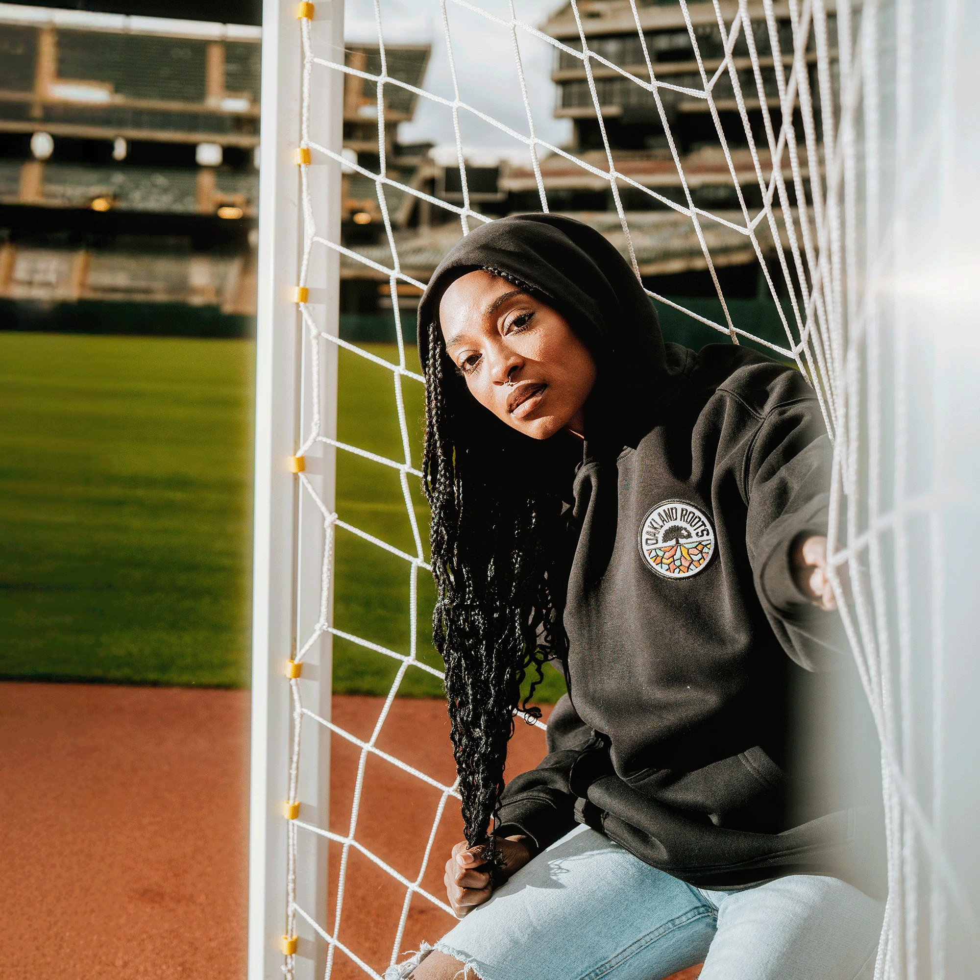 A woman with long braids wears the Oakland Roots SC Logo Premium Hoodie by Oakland Roots SC, paired with ripped jeans, sitting in a soccer goal under sunlight on an empty stadium field—capturing true Oakland Roots vibes.