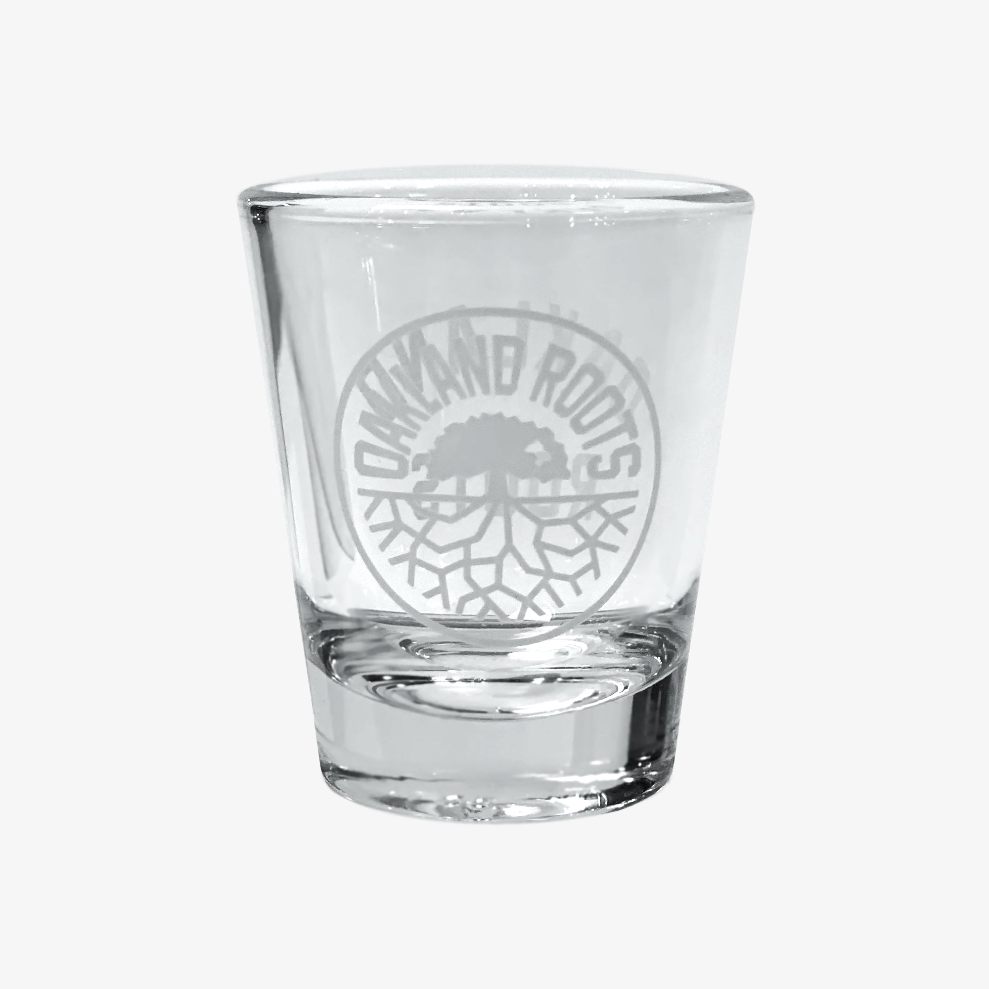 The Oakland Roots SC Shot Glass by Oakland Roots SC is a clear glass etched with the words "OAKLAND ROOTS" encircling a tree with spreading roots, capturing the spirit of the Oakland Roots Sports Club. The etching is perfectly centered on the front, set against a plain white background.