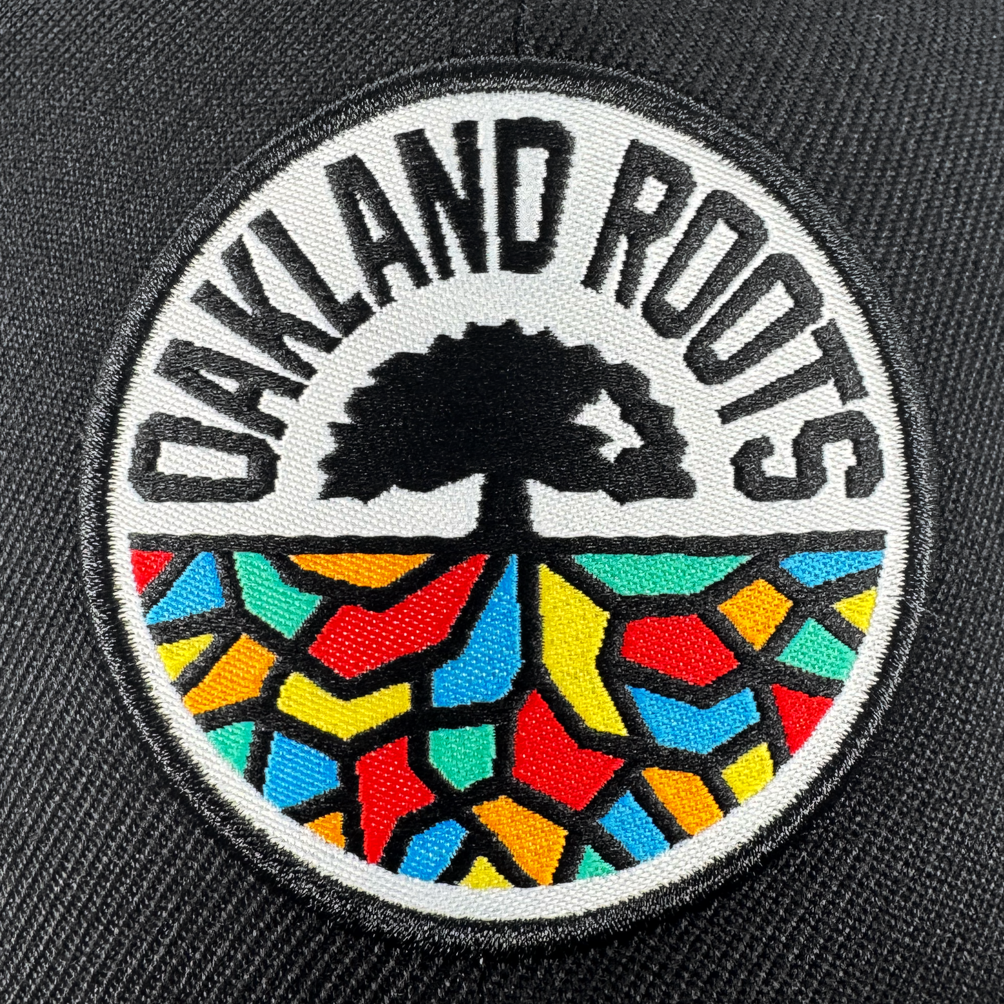 The Oakland Roots SC Structured Snapback features a circular patch with "OAKLAND ROOTS" around a black tree, colorful geometric roots below, and detailed stitching on dark fabric—celebrating the vibrancy of Oakland Roots SC.