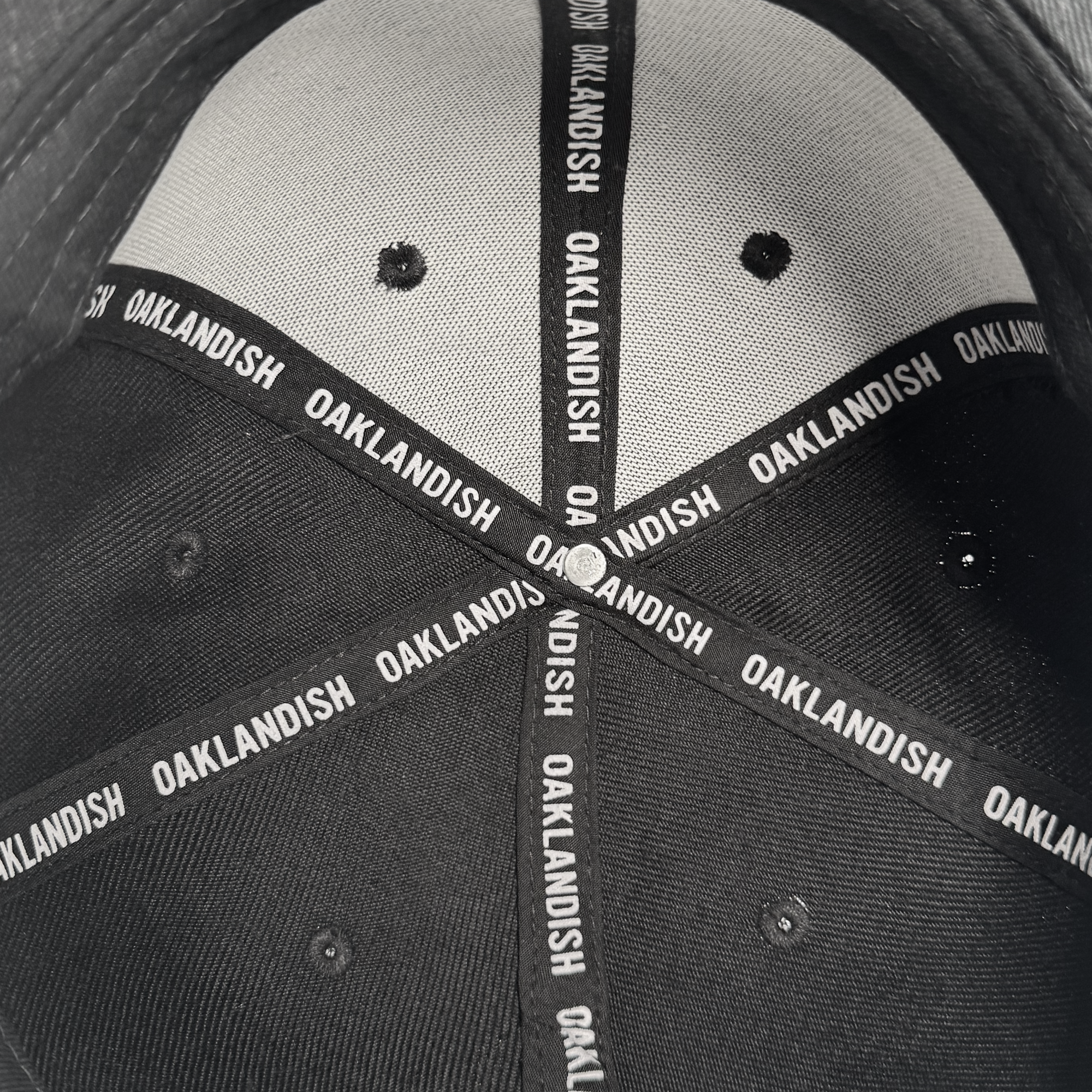 Close-up of the inside top of the Oakland Roots SC Structured Snapback by Oakland Roots SC. Black fabric strips with bold white "OAKLANDISH" lettering reinforce the seams, celebrating local sports spirit. Visible ventilation holes and stitching.