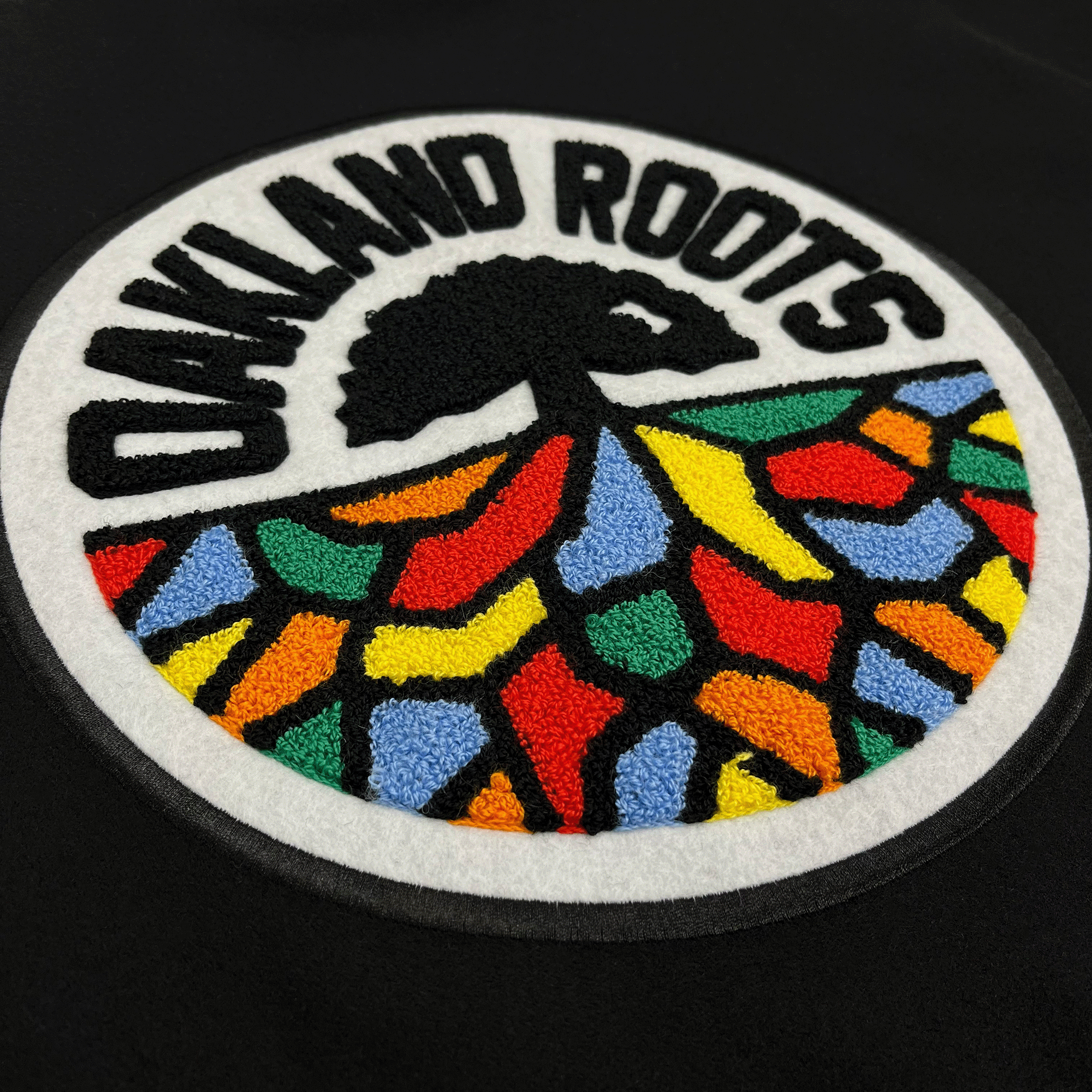 Side-angled close-up of large round full-color 3D Oakland Roots crest on the back of a black varsity jacket.