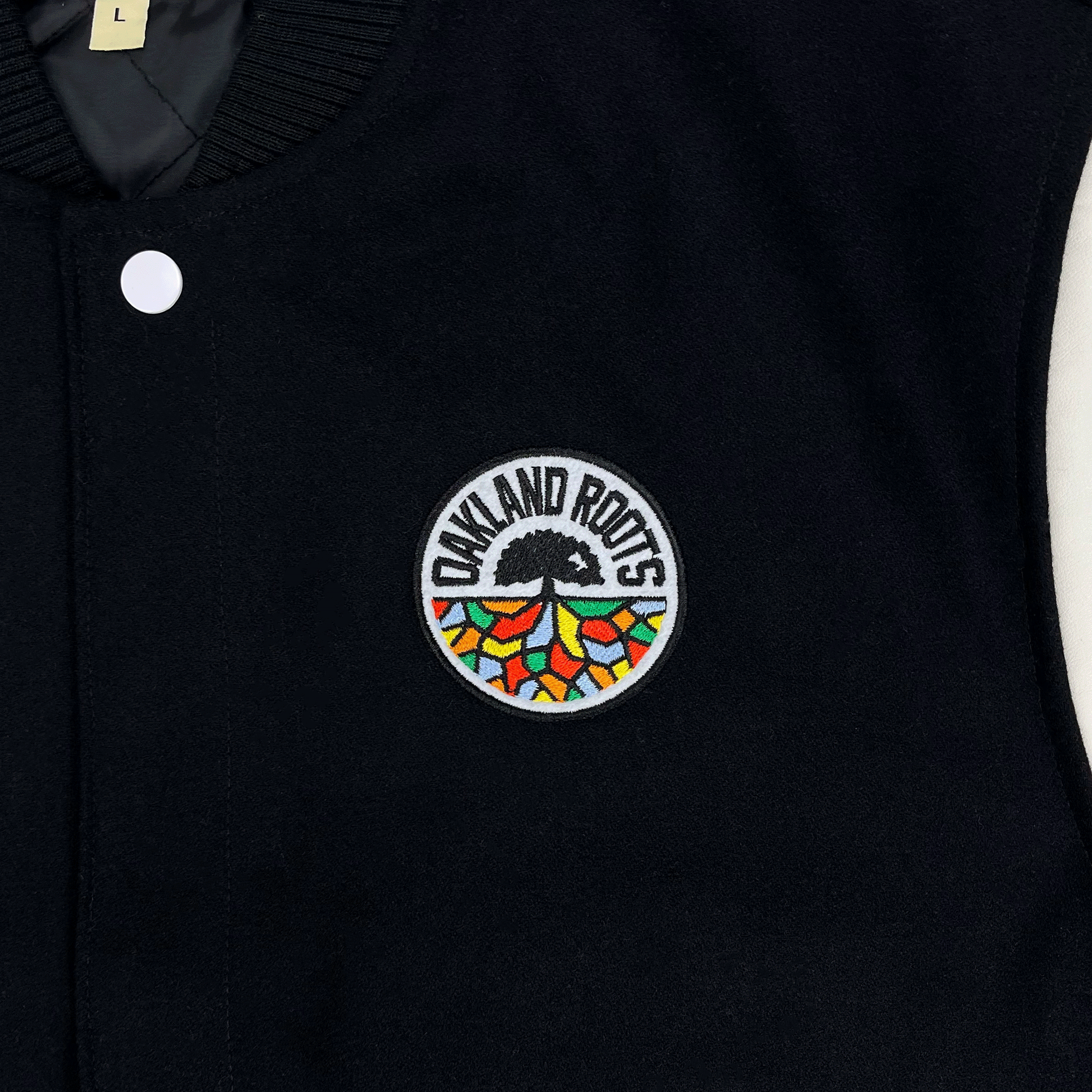 Close-up of full-color Oakland Roots logo on the left chest wearside of a black and white varsity jacket.