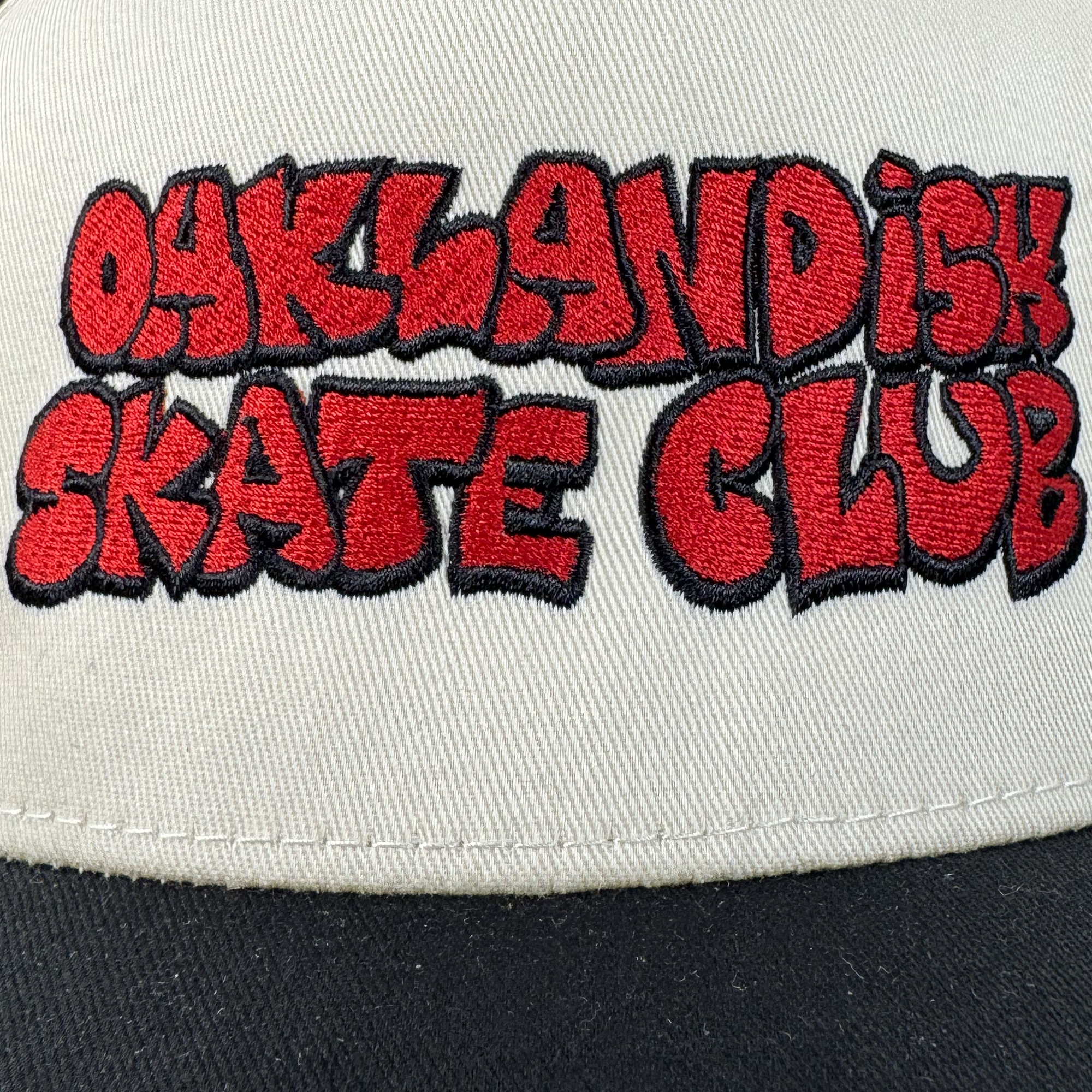 A close-up of the Oaklandish Skate Club 2-Tone Hat by Oaklandish, featuring bold red and black graffiti-style embroidery on beige with a black brim—an authentic nod to skateboard culture's streetwear edge.