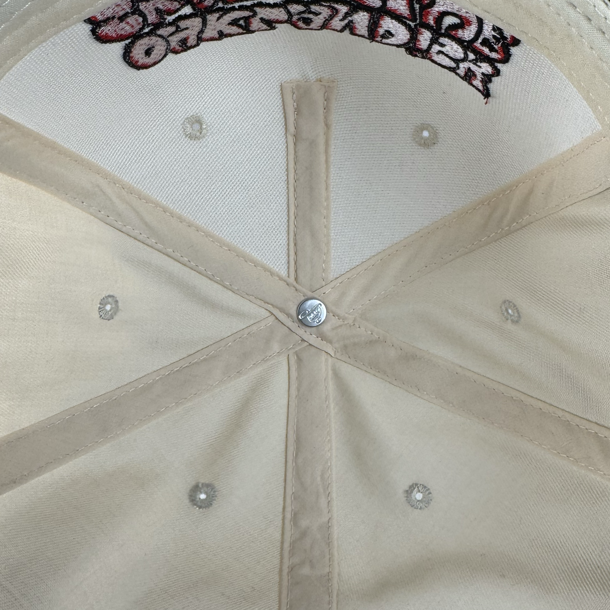 Close-up of the inside of an Oaklandish Skate Club 2-Tone Hat, showing stitched seams meeting at a central silver button. Small embroidered circles decorate each panel, with part of the pink and black Oaklandish logo visible at the top edge.