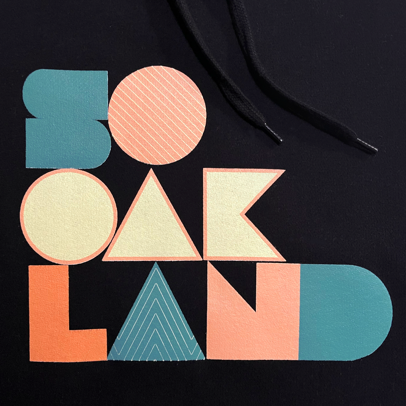 Detailed close-up of large multi-color SOOAKLAND graphic wordmark on the back of a black pullover hoodie.