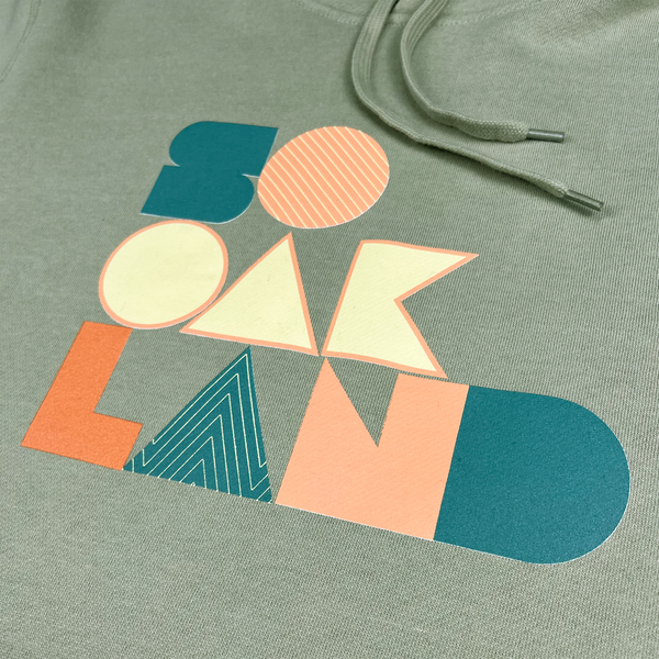 Detailed close-up of large multi-color SOOAKLAND graphic wordmark on the back of a pistachio pullover hoodie.