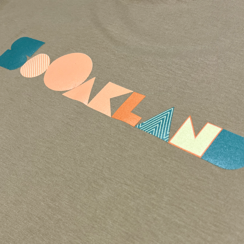 Detailed close-up of full-color SOOAKLAND graphic wordmark on the back of a sand-colored long-sleeved t-shirt.