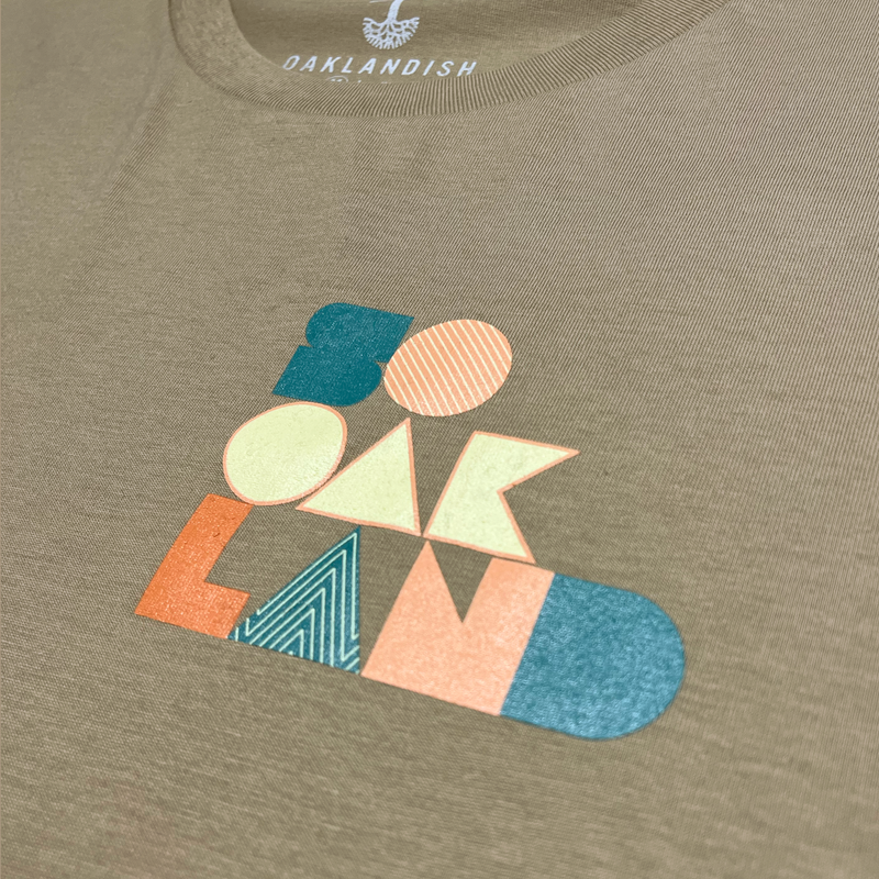 Detailed close-up of full-color SOOAKLAND graphic wordmark on the chest of a sand-colored long-sleeved t-shirt.