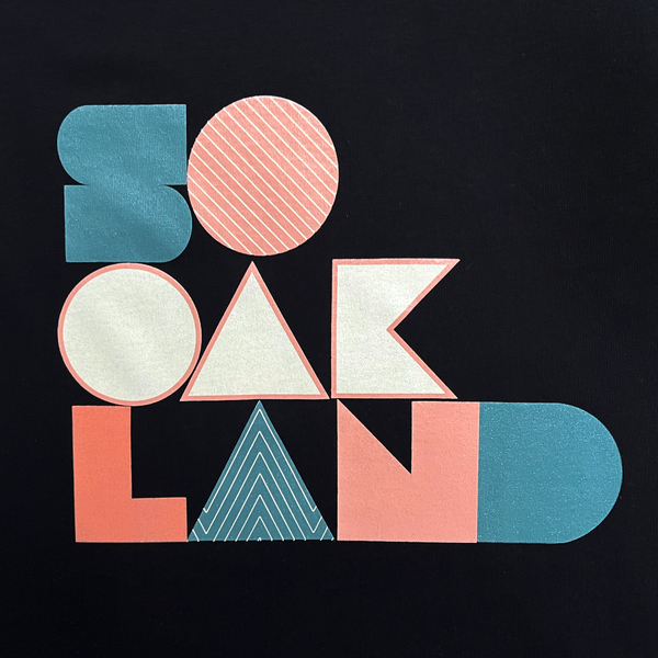 Close-up of SoOakland full-color wordmark on the front chest of a black women’s cut t-shirt.