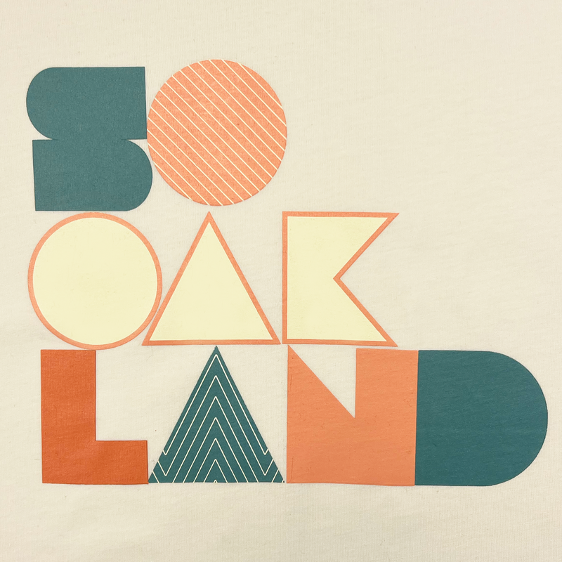 Close-up of SoOakland full-color wordmark on the front chest of a natural cotton colored t-shirt.