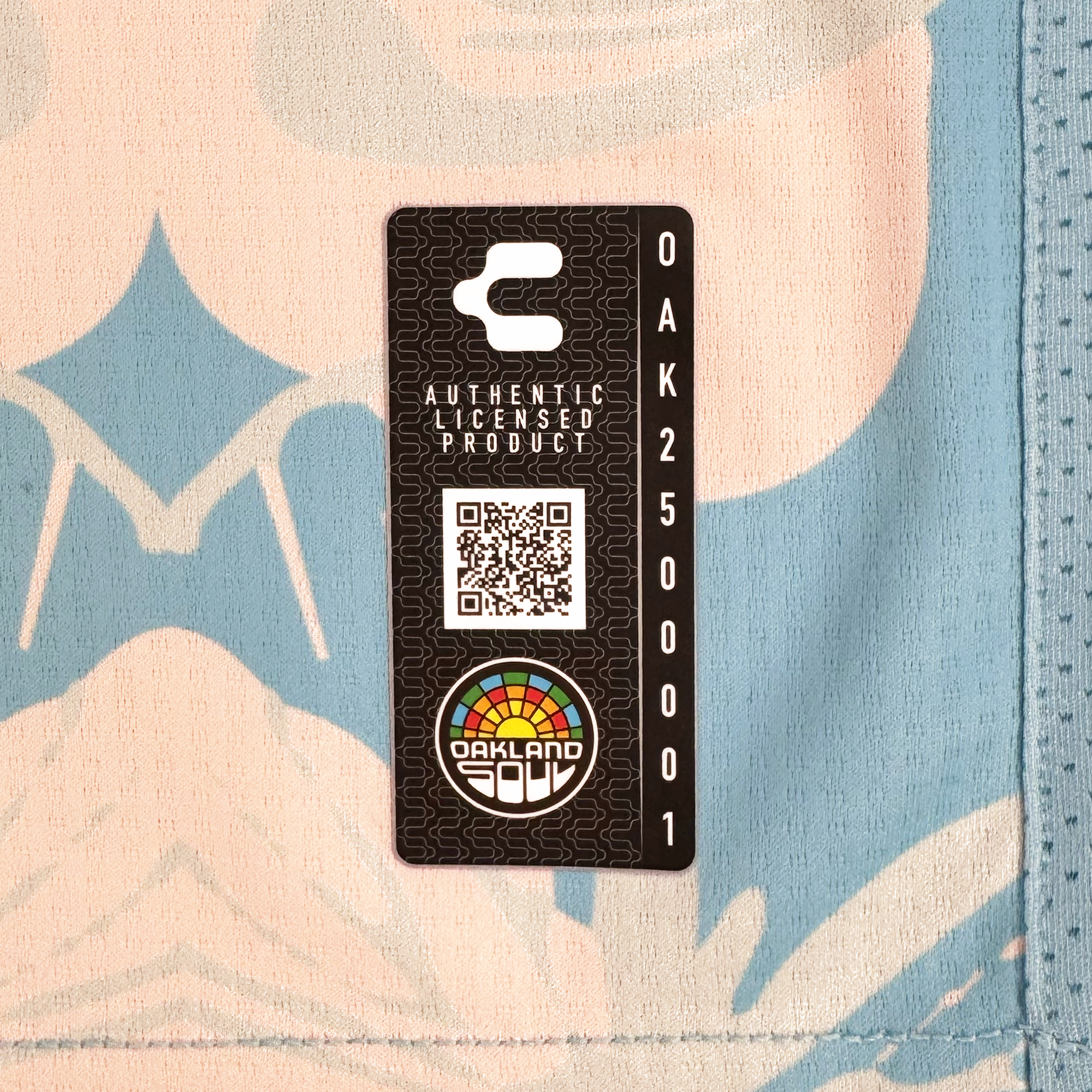 A black rectangular tag stitched to the Custom Butterfly Effect Kit's floral tie-dye fabric displays a QR code, “AUTHENTIC LICENSED PRODUCT,” “OAK 250001,” a colorful Oakland Soul logo, and a white stylized “C.”.