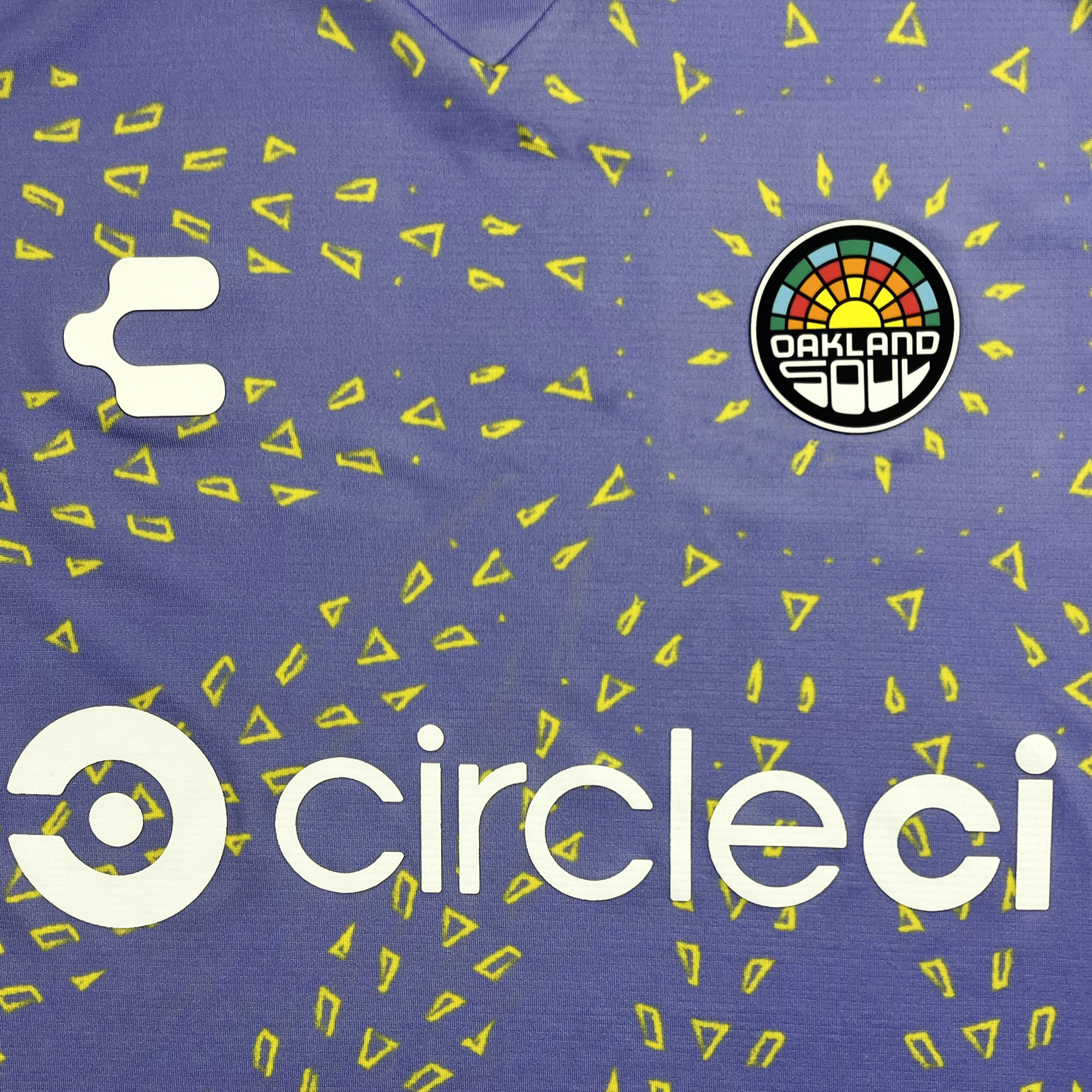 A close-up of the Custom Unisex Oakland Soul Second Kit ft. CircleCI sports jersey, showcasing a colorful, circular Oakland Soul logo on the upper right. Scattered yellow triangle patterns embellish the purple fabric. Below the logo, white text spells "circleci" alongside its stylized letter "C." The tie-dye design adds a vibrant touch to this athletic wear from Oakland Soul.