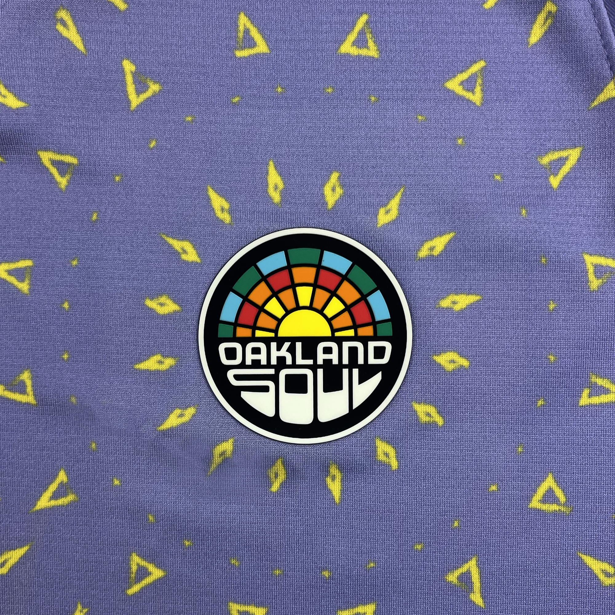 A vibrant and energetic design on purple fabric adorned with yellow triangular patterns radiating outward from the "Oakland Soul" logo, which features a stylized sunset with rays in blue, orange, and green. This design is reminiscent of a tie-dye pattern and is part of the Custom Unisex Oakland Soul Second Kit ft. CircleCI by Oakland Soul.