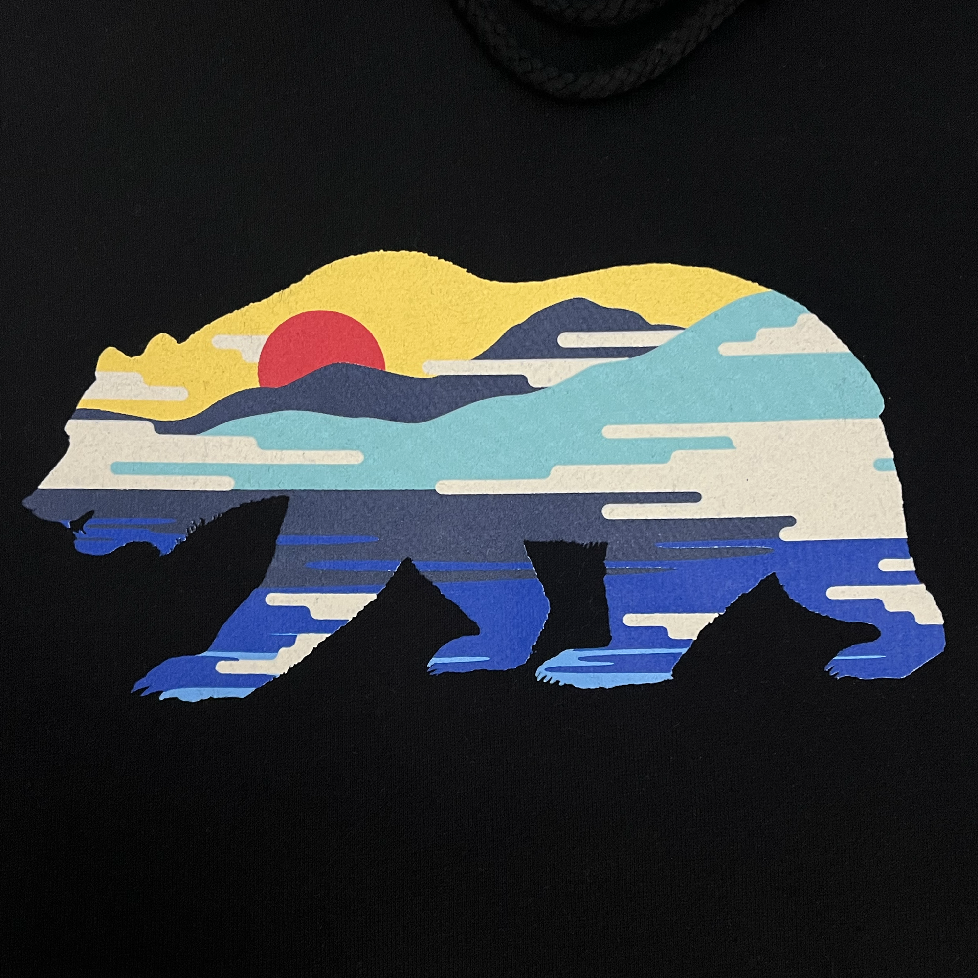 The Sunset Bear Hoodie by Never Elsewhere features an illustration of a California grizzly bear silhouette filled with a vibrant landscape scene. Within the silhouette, a red sun sets or rises over blue mountains and water, beneath a sky scattered with clouds. The black background emphasizes the striking colors within the bear shape. This design is available in men's sizing on a pullover hoodie.