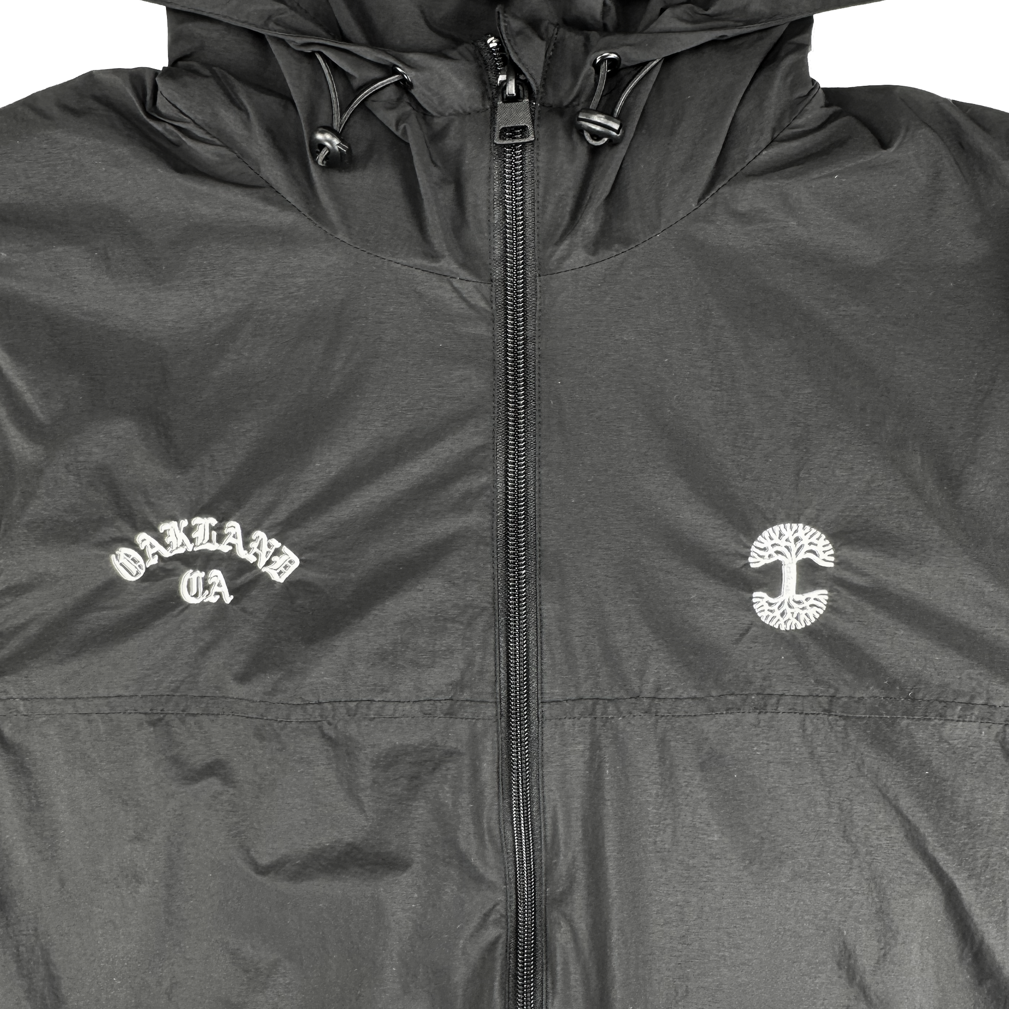The Susurrus Track Top by Oaklandish is a black nylon zip-up jacket with a subtle sheen. "OAKLAND CA" is printed in white Old English on the left chest, with a circular tree logo on the right—a standout men's outerwear piece.