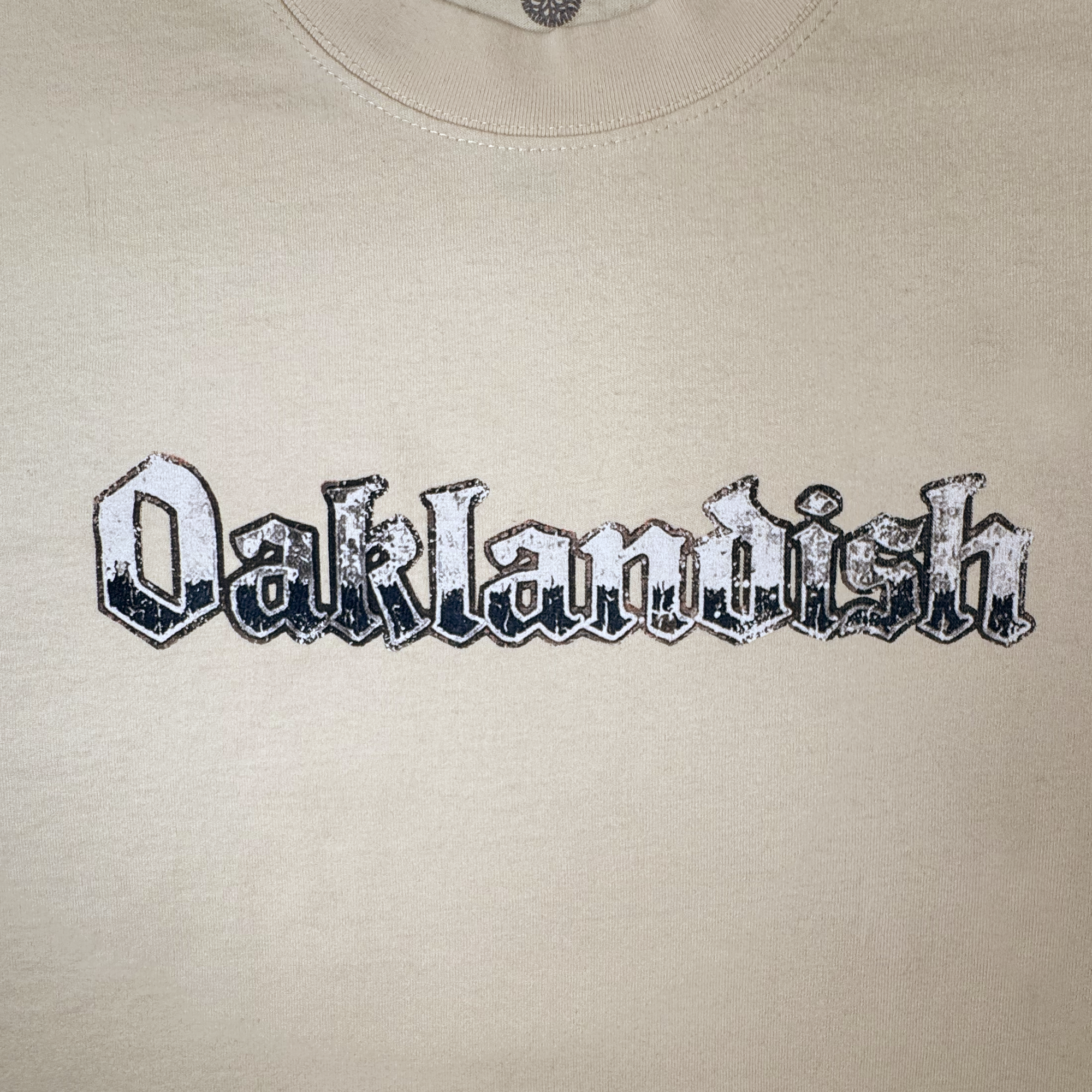 Introducing the Oaklandish Swoop Tee: a classic fit beige t-shirt featuring the word “Oaklandish” boldly printed across the chest in a distressed blackletter-style font. The weathered, vintage-effect text—rendered in white and black—complements the shirt’s soft, heavy-weight cotton fabric with its subtly textured feel.