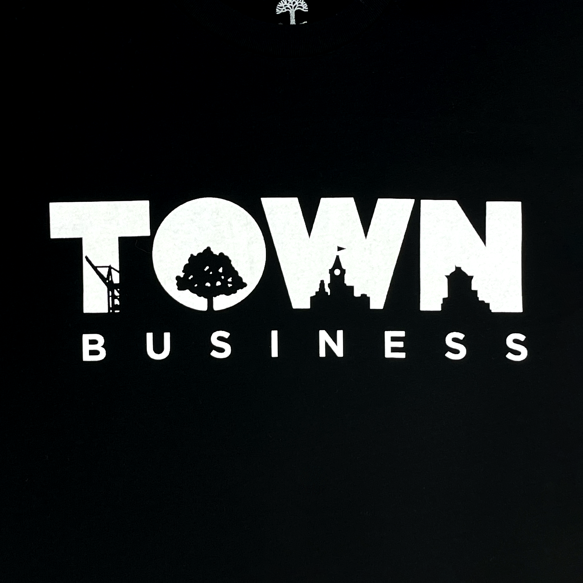 The image showcases the Town Business 2024 Tee by Oaklandish. It features a black background with white typography that reads "TOWN BUSINESS," where the letters in "TOWN" creatively incorporate various silhouettes: a crane in the "T," an Oakland tree in the "O," a clock tower in the "W," and a building in the "N.
