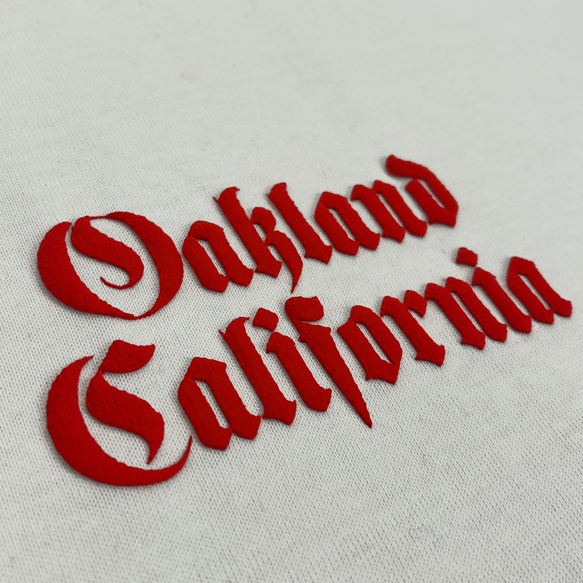 The Oaklandish Town Rose Heavy Tee features stylized text in a gothic font, embroidered in red thread on heavyweight combed cotton fabric that reads "Oakland California." The text is split into two lines with "Oakland" on top and "California" below it. The embroidery is bold and slightly raised, ideal for a boxy fit t-shirt designed for men.